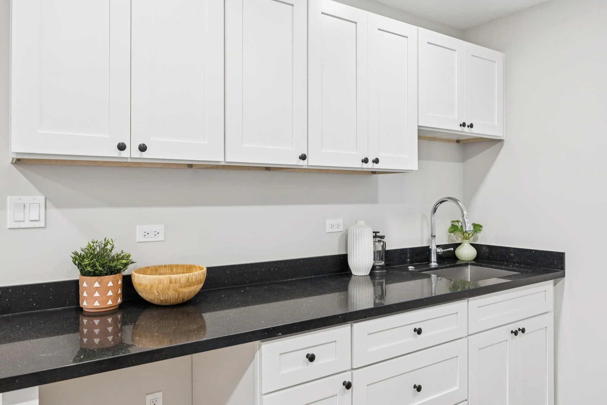 White kitchen cabinets with black knobs, black granite countertop, a stainless steel sink with a curved faucet, and decor items including potted plants and bowls.