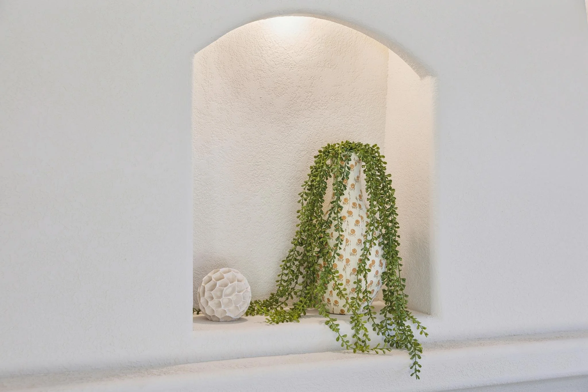 A white wall with a textured alcove containing a tall, leafy green plant in a white pot with a decorative pattern, and a white geometric lamp in the foreground.