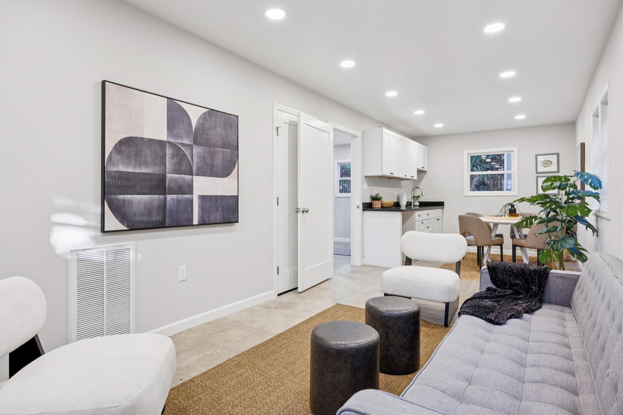A living room with white walls and ceiling, recessed lighting, a gray sofa with a black throw, white armchairs, black ottomans, a large wall art piece with abstract shapes, a small round table with a potted plant, and a view into a dining area with w