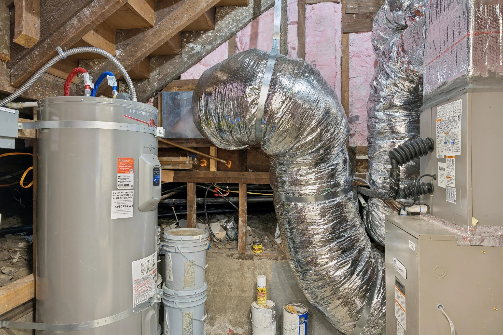 Construction or renovation room with exposed insulation and ductwork. Two water heaters are visible, along with various pipes, wiring, and construction materials on the floor.