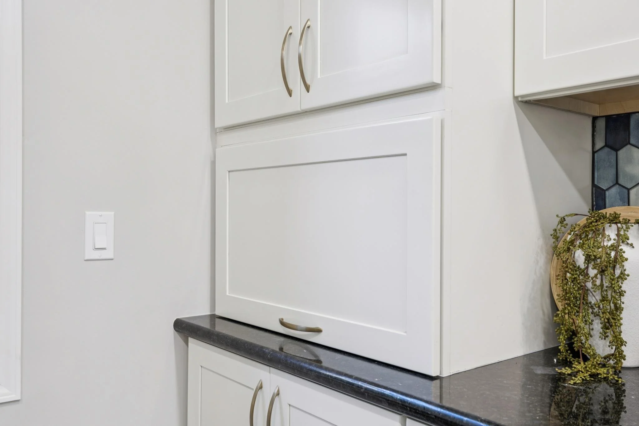 A close-up of a kitchen cabinet with silver handles, part of a white kitchen wall, a black countertop, and a decorative plant in a basket.