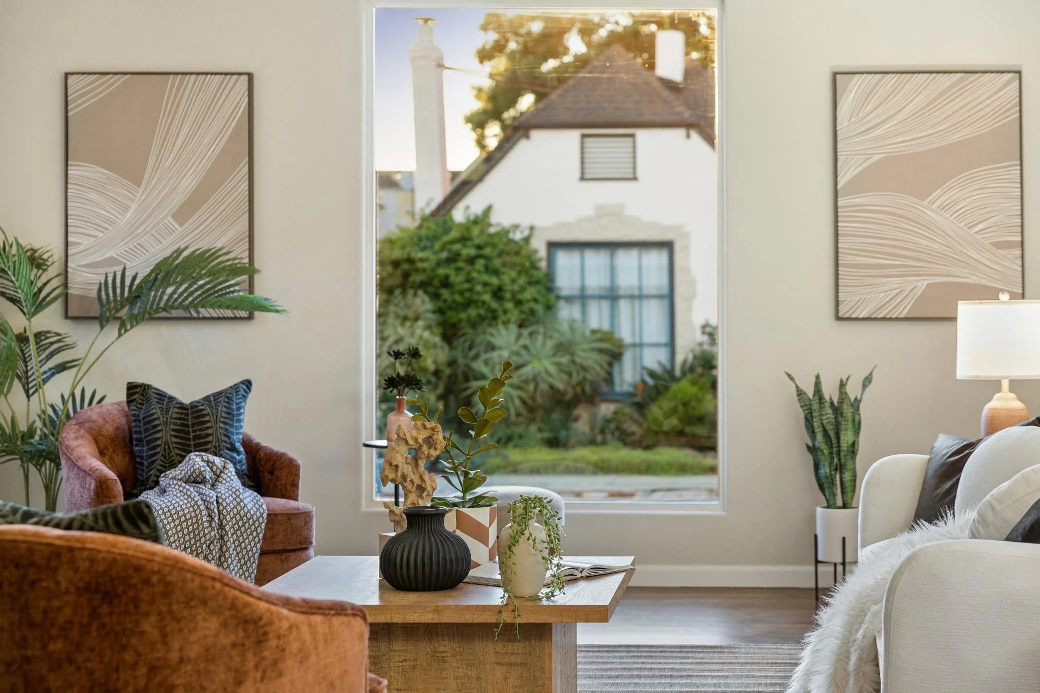 Living room with large window, potted plants, abstract wall art, velvet armchairs, a coffee table with decorative items, and a mix of neutral and bold-colored furnishings.