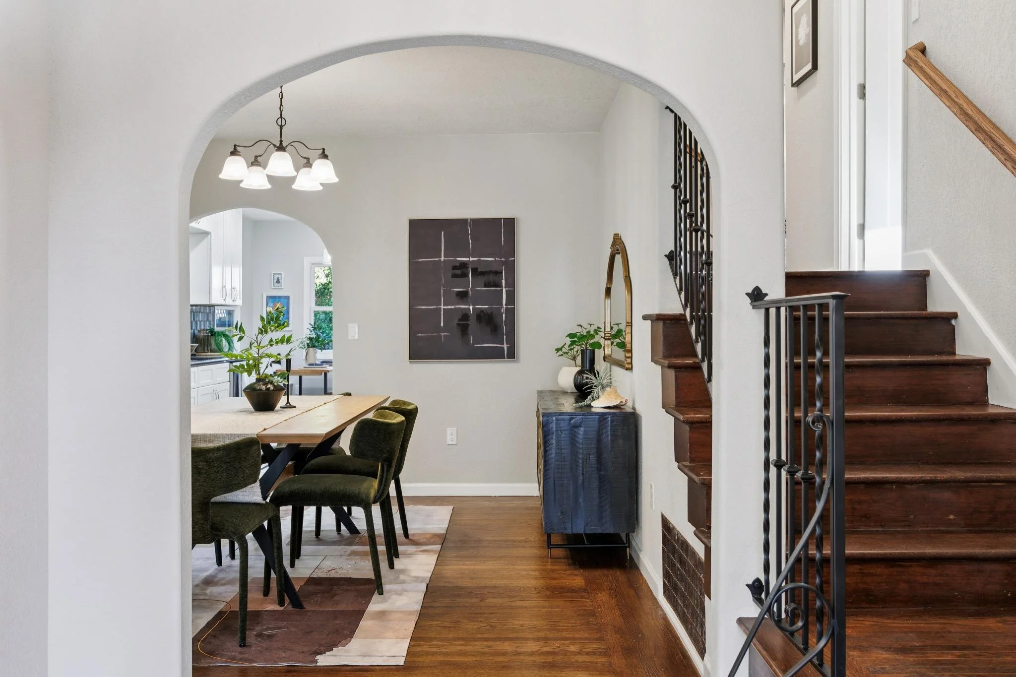 Interior view of a house focusing on a staircase with dark wood steps and an ornate black railing, leading to the upper floor. The wall beside the staircase has a framed picture and a mirror. To the left, a doorway opens into a dining area with a tab