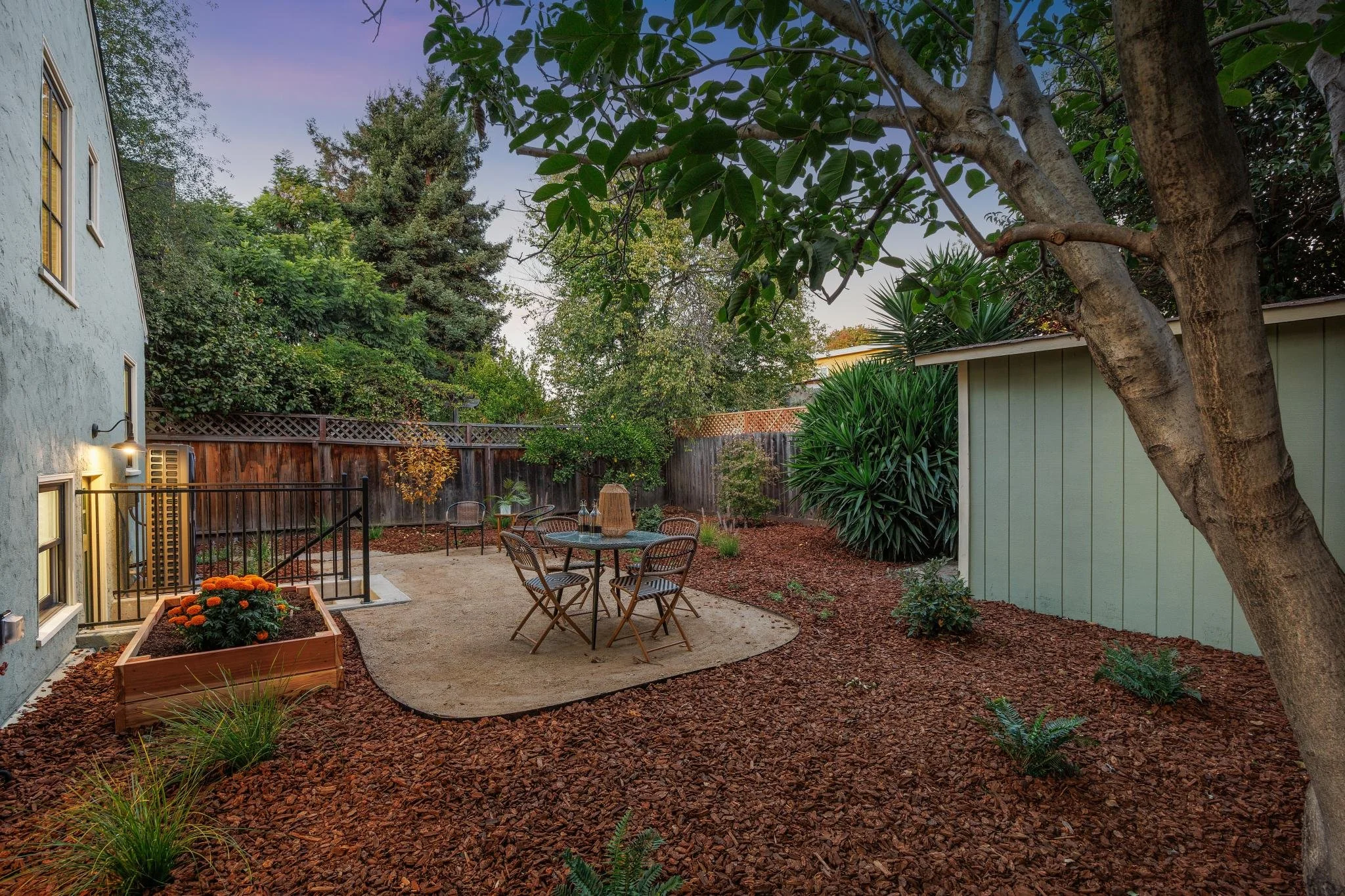 A backyard patio with a table and four chairs, surrounded by trees and bushes, with a wooden fence in the background and sunset lighting.