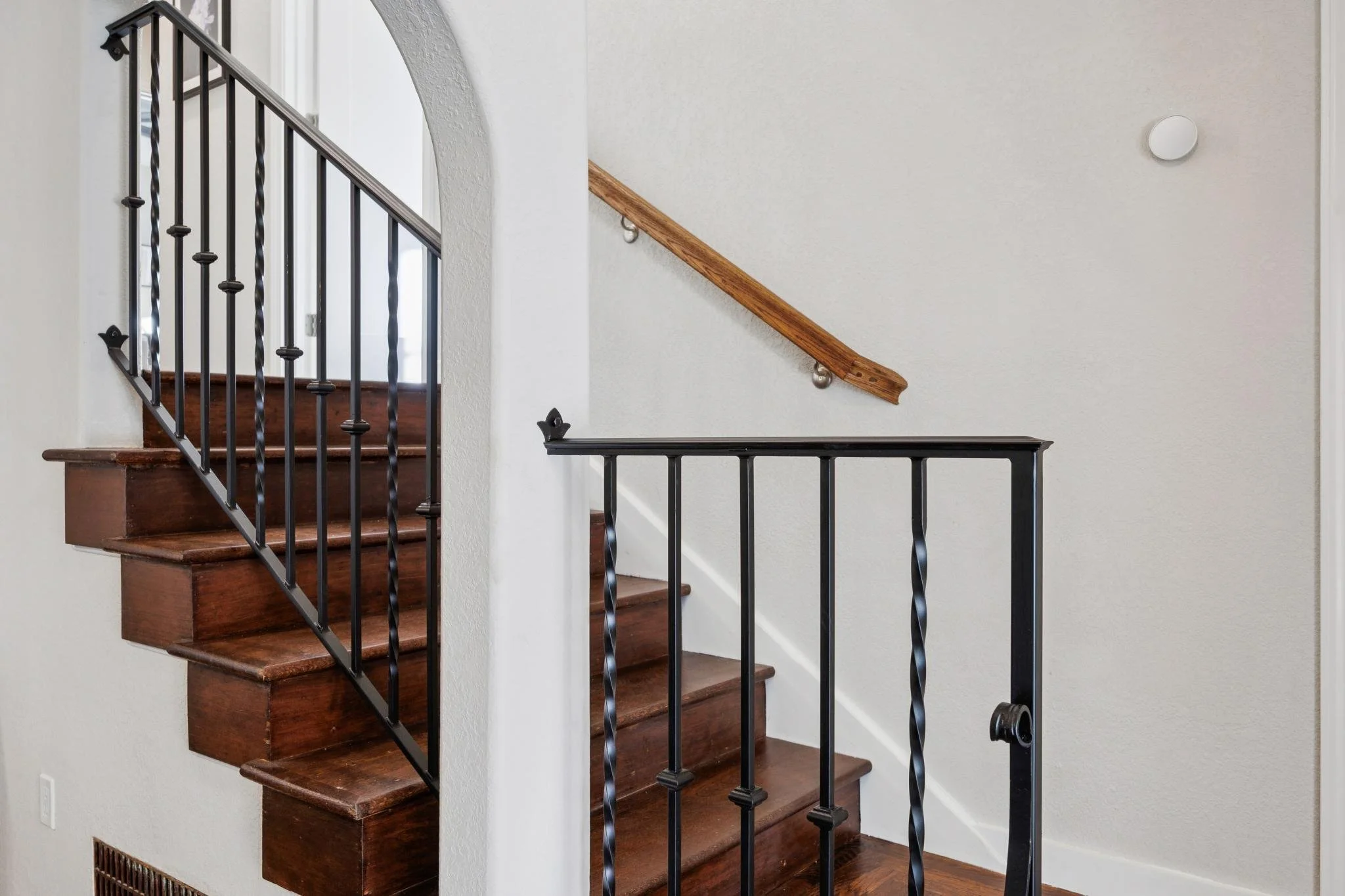 Interior view of a staircase with dark wooden steps, black wrought iron balusters, and a wooden handrail, with white walls surrounding it.