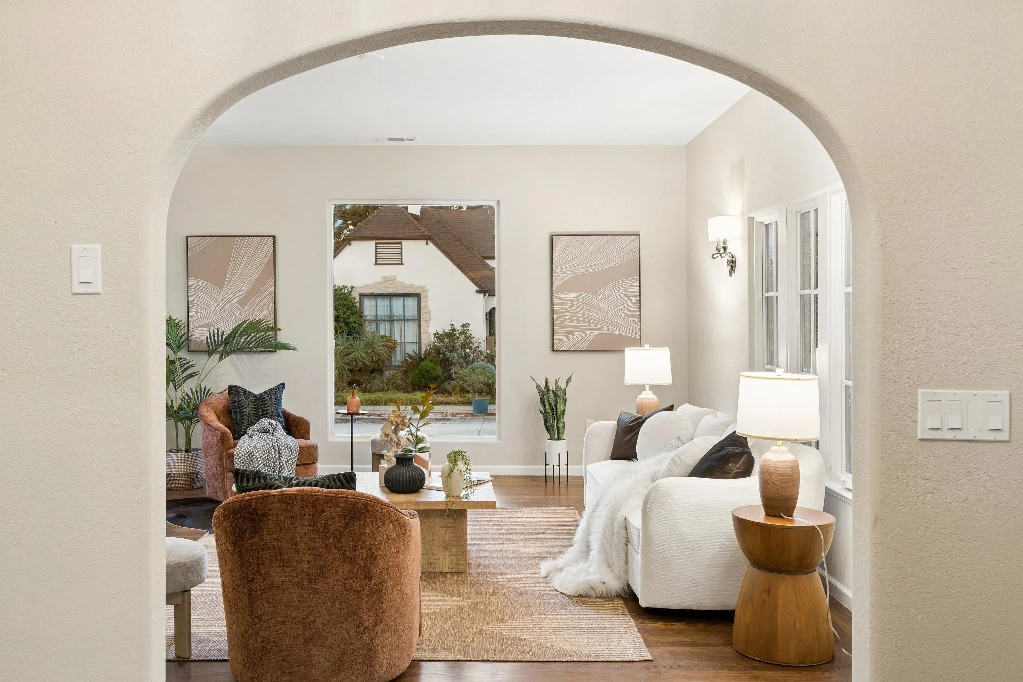 A living room viewed through an arched doorway. It contains white and brown sofas, a wooden coffee table, various houseplants, and framed art on the wall. A large window reveals a view of neighboring houses and greenery outside.