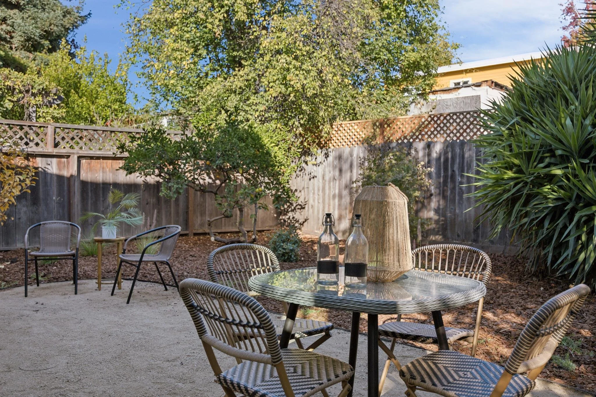A backyard patio area with wicker chairs surrounding a glass table, bottle lanterns, and a tree with green and yellow leaves, enclosed by a wooden fence and lush bushes.