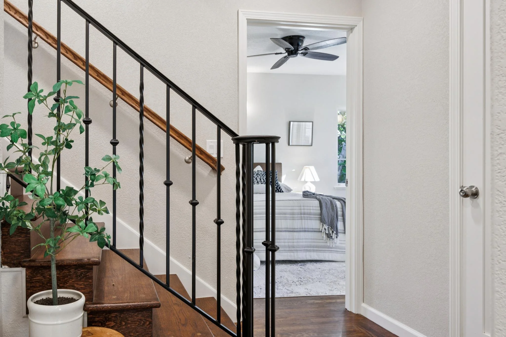 Interior of a house showing a staircase with black metal railing, a potted plant, and a doorway leading to a bedroom with a bed, pillow, blanket, lamp, and ceiling fan.