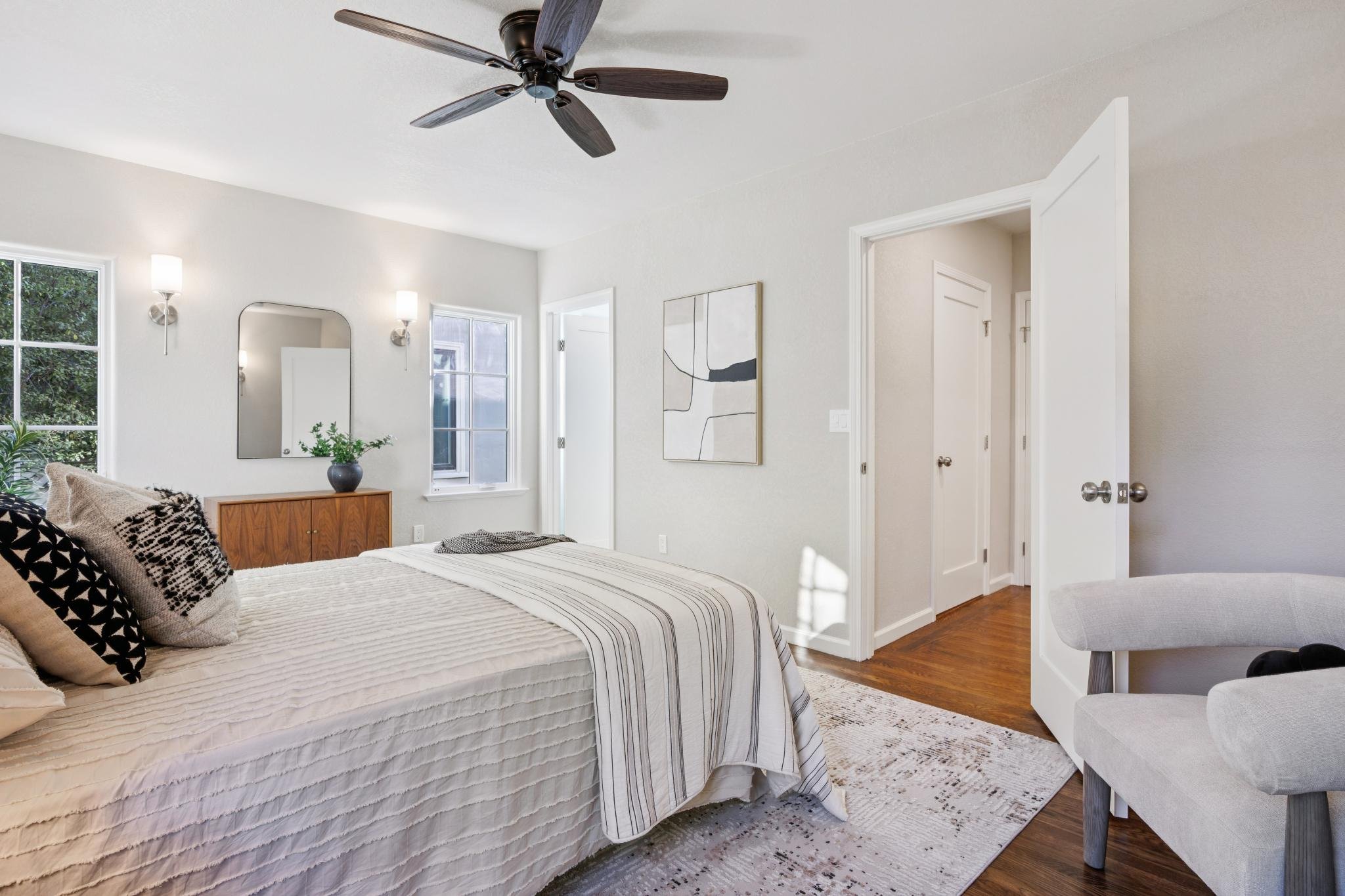 A bedroom with a bed covered in a cream-colored quilt and decorative pillows, a wooden dresser with a plant, and a wall mirror. There are two wall-mounted lights, windows allowing natural light, and artwork on the wall. A ceiling fan with dark blades