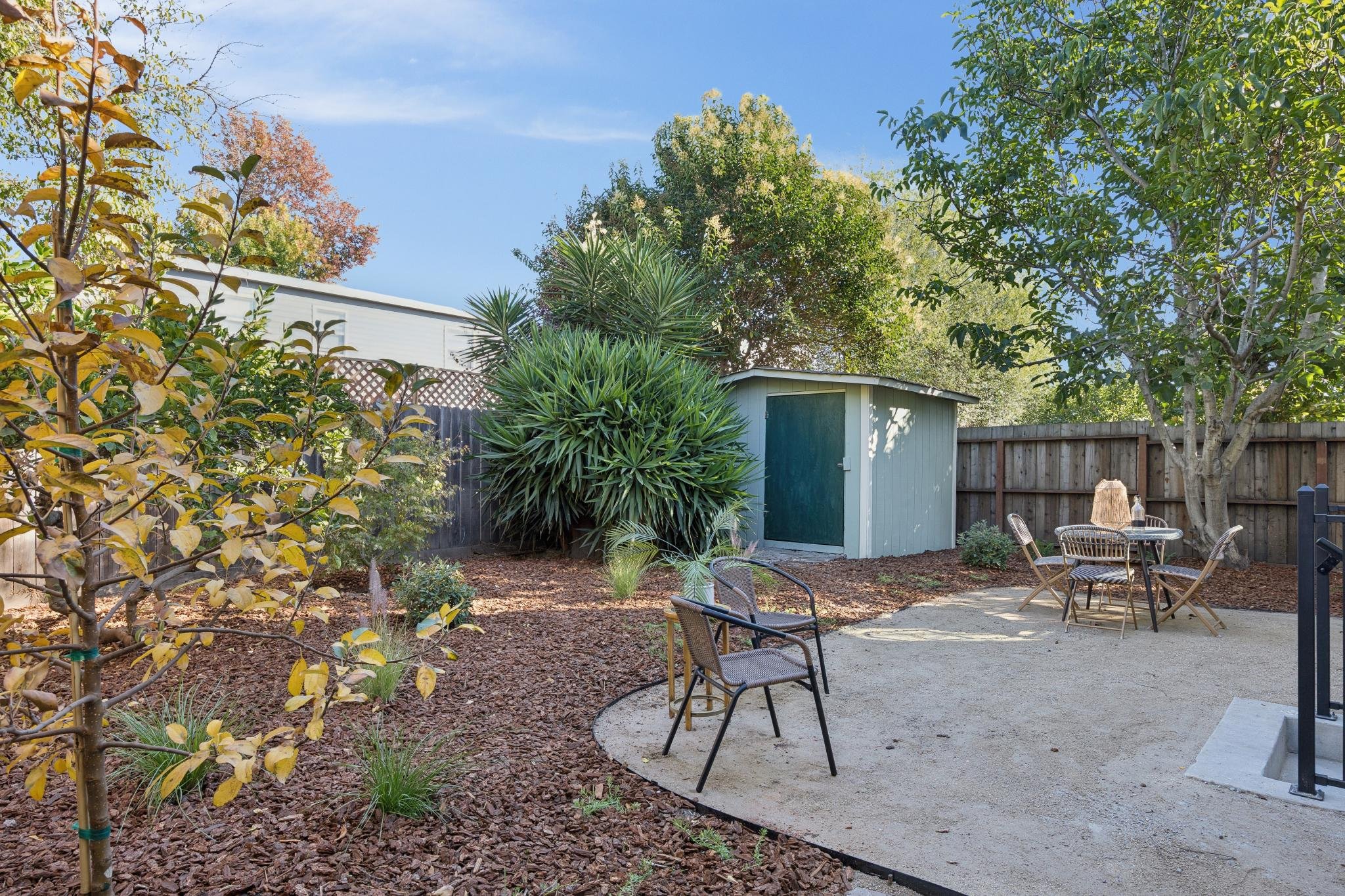 A backyard garden with a small shed, various trees, and outdoor seating including tables and chairs. The ground is covered with mulch and a concrete patio area.