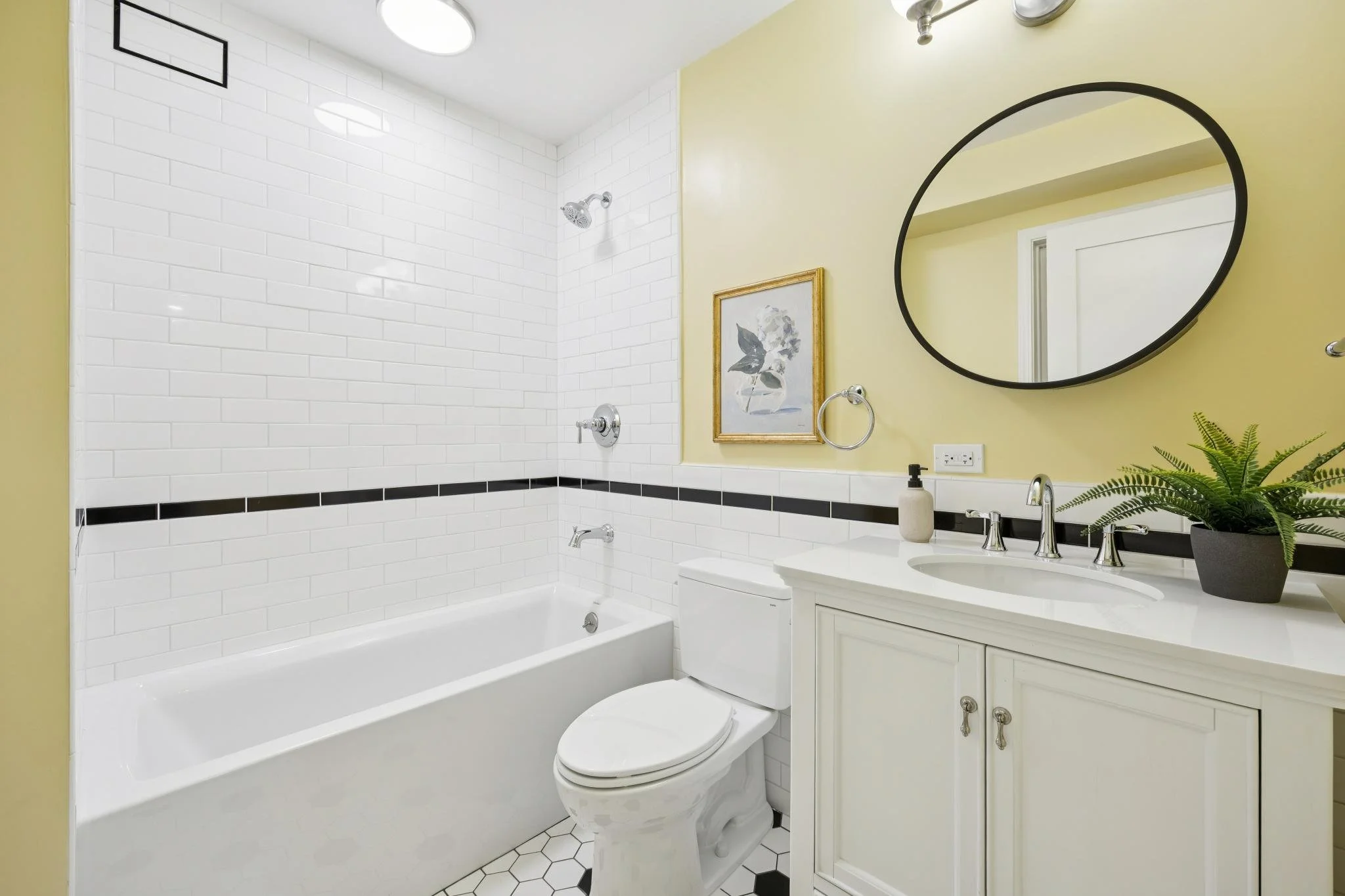 Bathroom with white subway tile on the walls, a bathtub with a showerhead, a white vanity with a sink, a round mirror, and a yellow wall with artwork and a plant.