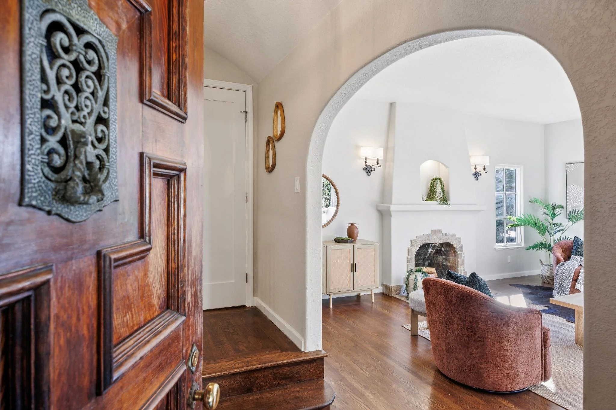 View through a doorway into a living room with white walls, a fireplace, a window, and modern wall-mounted lights. The room has a cozy, contemporary style with plants and comfortable furniture.