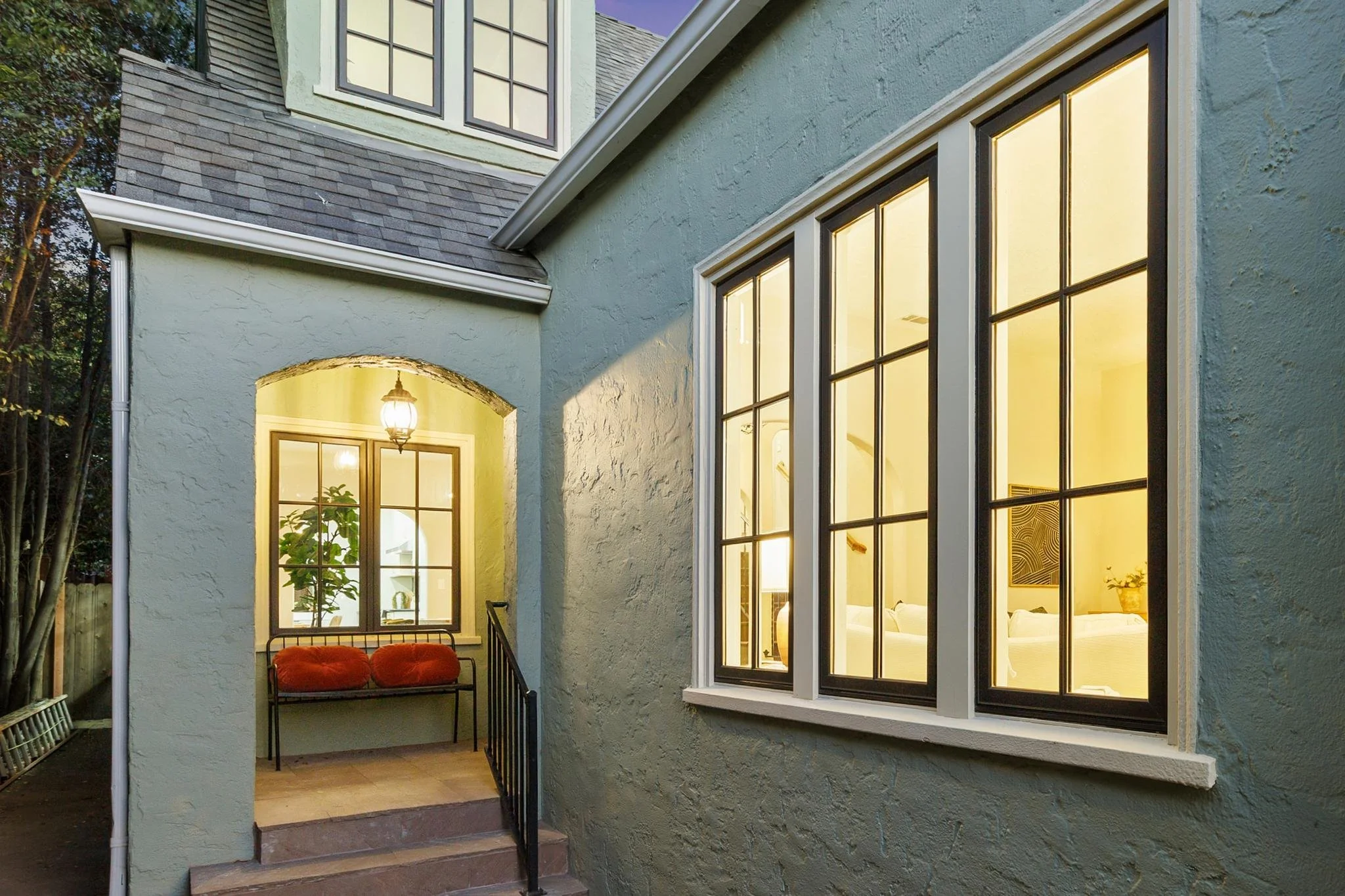 A house exterior with large window panes showing a warmly lit interior, a front porch with a bench with red cushions, and an outdoor lantern hanging near the doorway. The house has textured light-colored walls and a sloped roof.