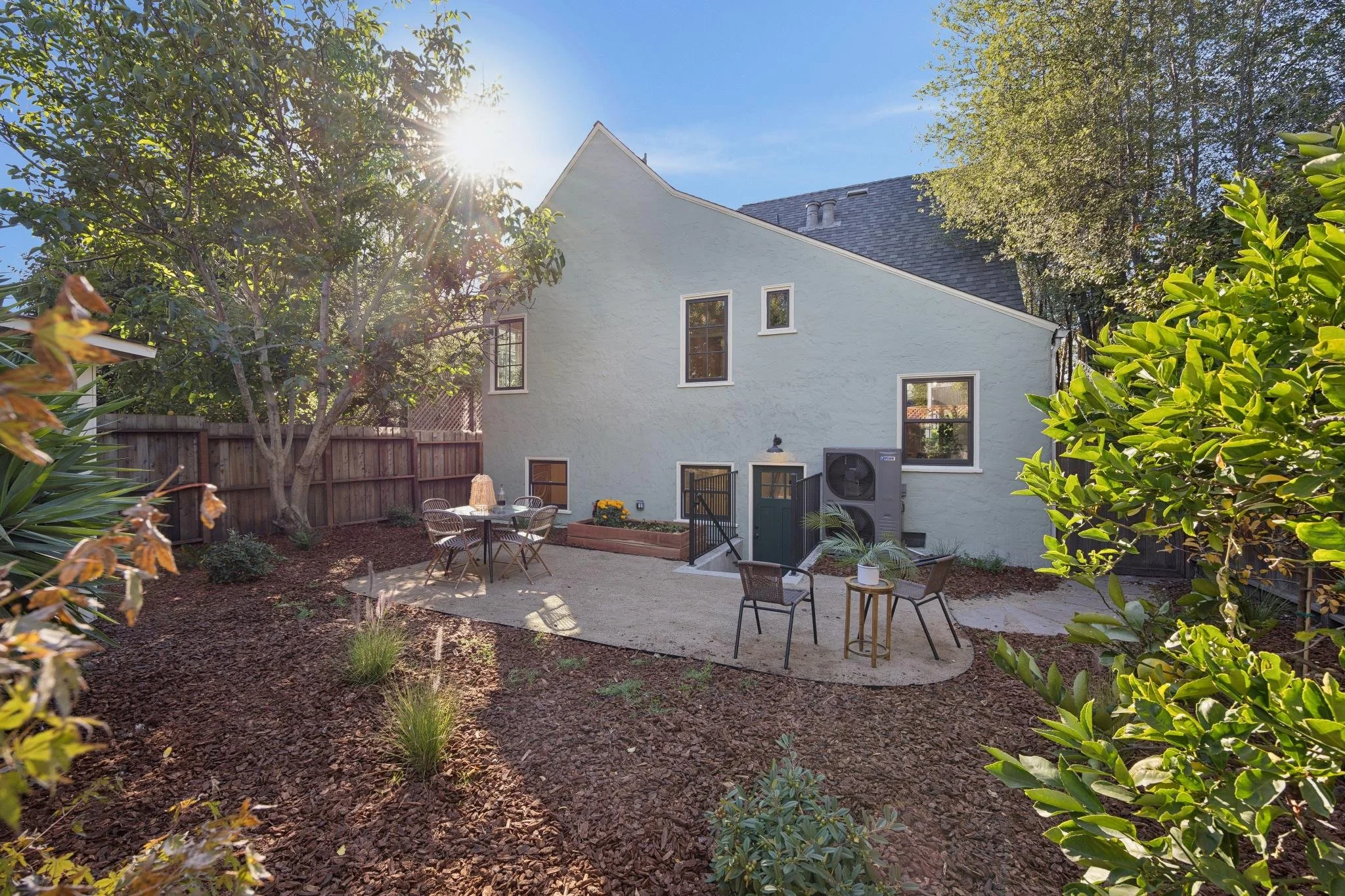 Backyard with patio, outdoor furniture, trees, and a light-colored house with multiple windows, under a clear blue sky with the sun shining through the trees.