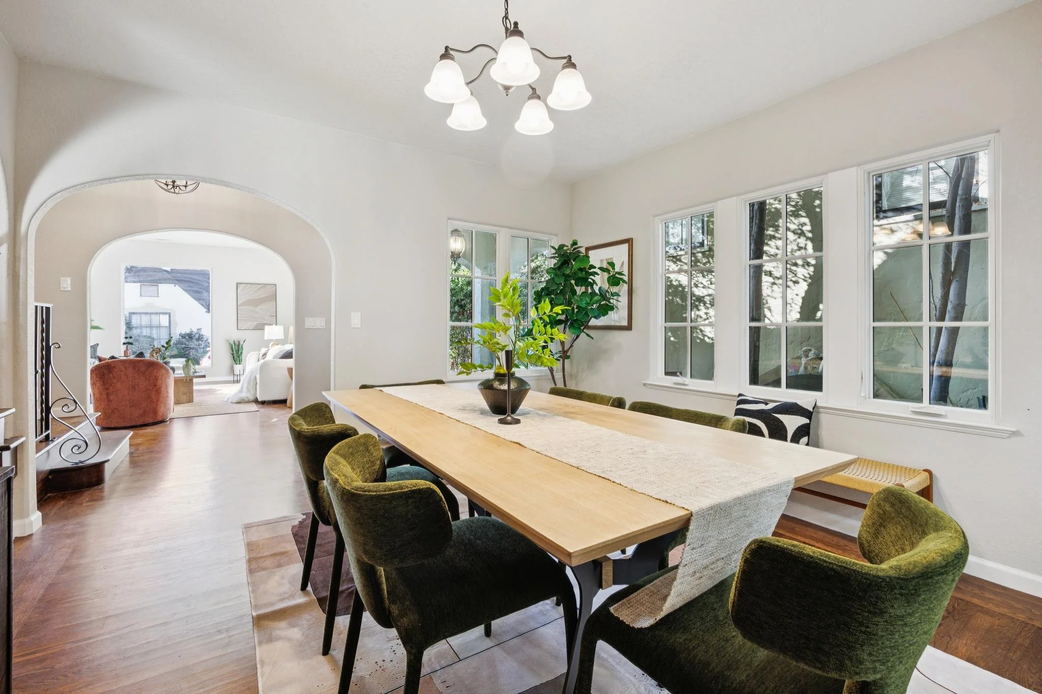 A dining room with a large wooden table and green upholstered chairs, surrounded by large windows showing trees outside, with a chandelier hanging from the ceiling.