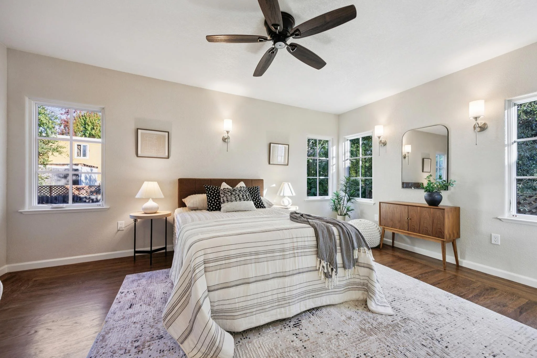 A bedroom with a bed, striped bedding, a wooden dresser with a plant, windows, and wall-mounted lights, with a ceiling fan overhead