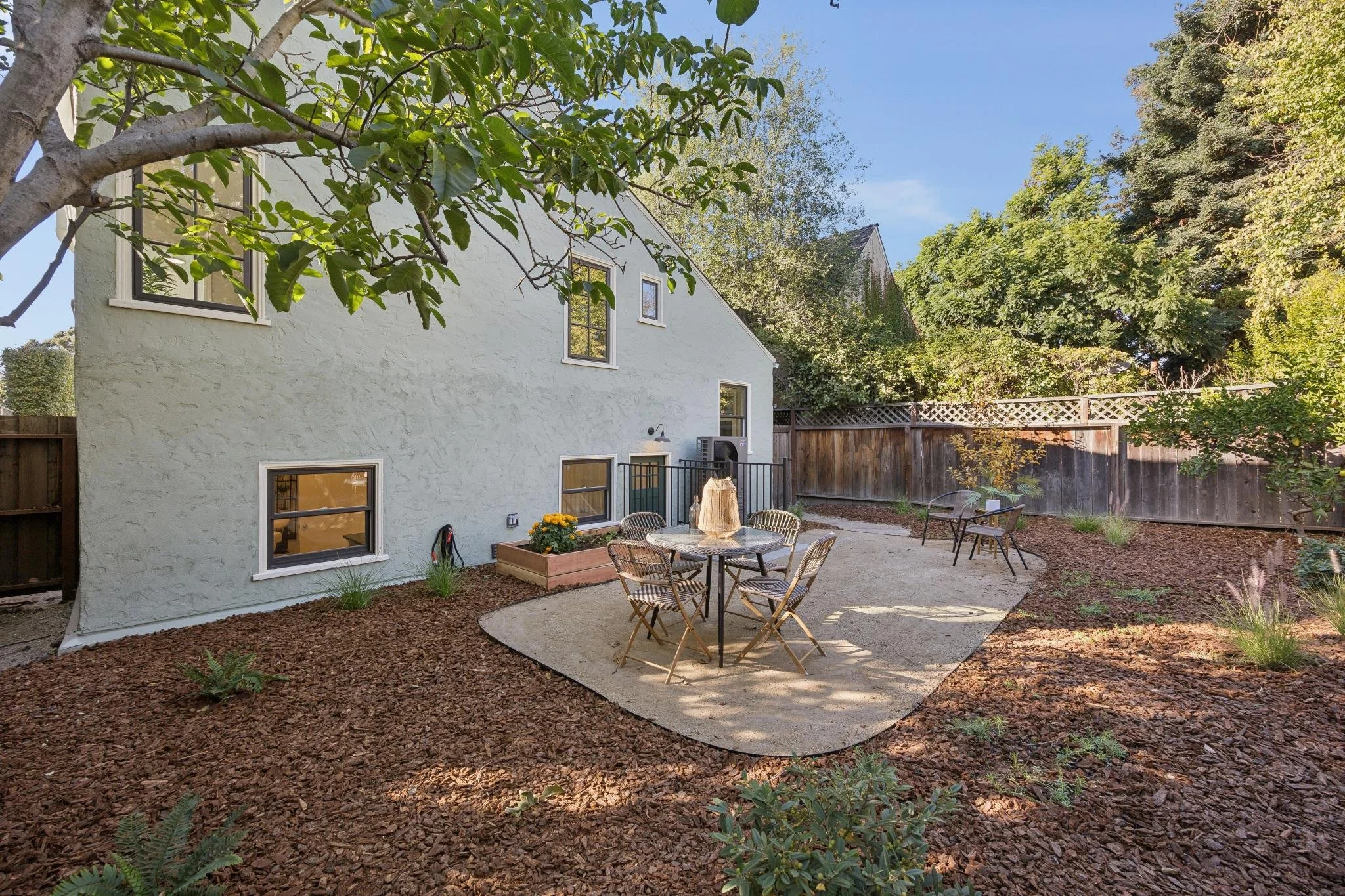 A backyard patio area with a white two-story house in the background, surrounded by green trees and a wooden fence. The patio has a concrete surface with an outdoor dining set, including a round table and several chairs, some with striped cushions. T