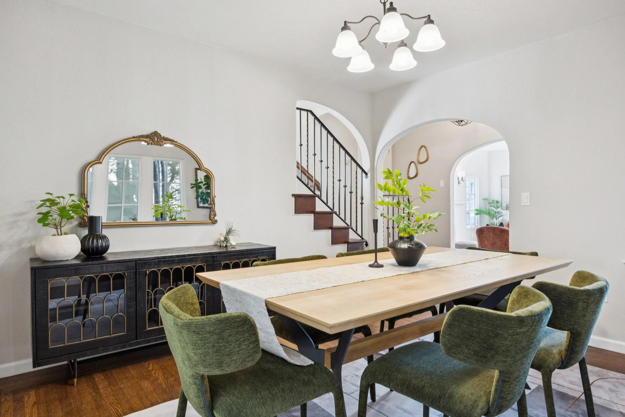 A dining area with a wooden table surrounded by green upholstered chairs, decor on a black sideboard with a mirror, and a staircase in the background.