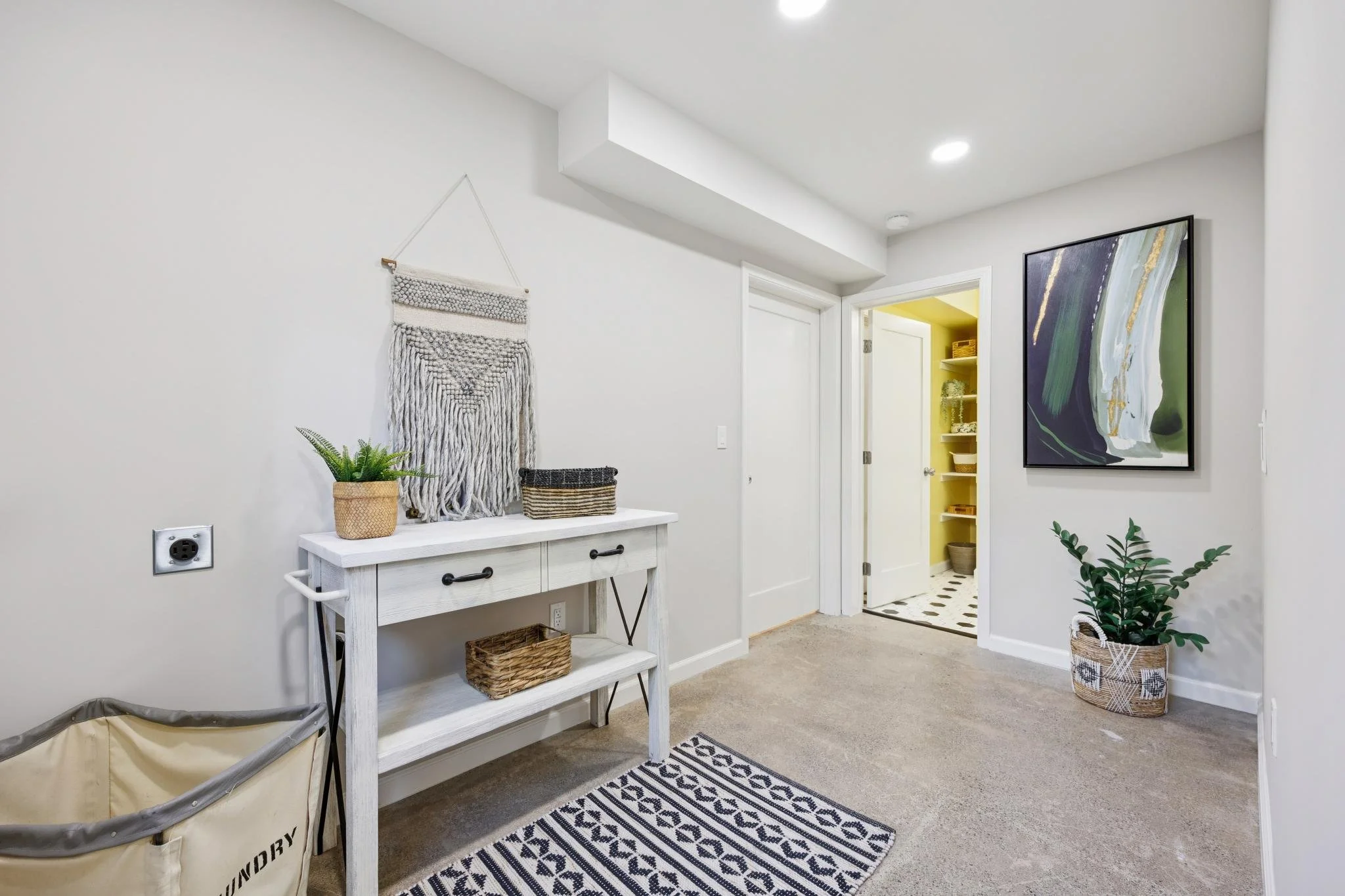 Entryway with white walls, a black and white patterned rug, a white console table with baskets, a potted plant, and wall decor with a woven wall hanging. There is a doorway leading to a yellow room with shelves and a bathroom with black and white til