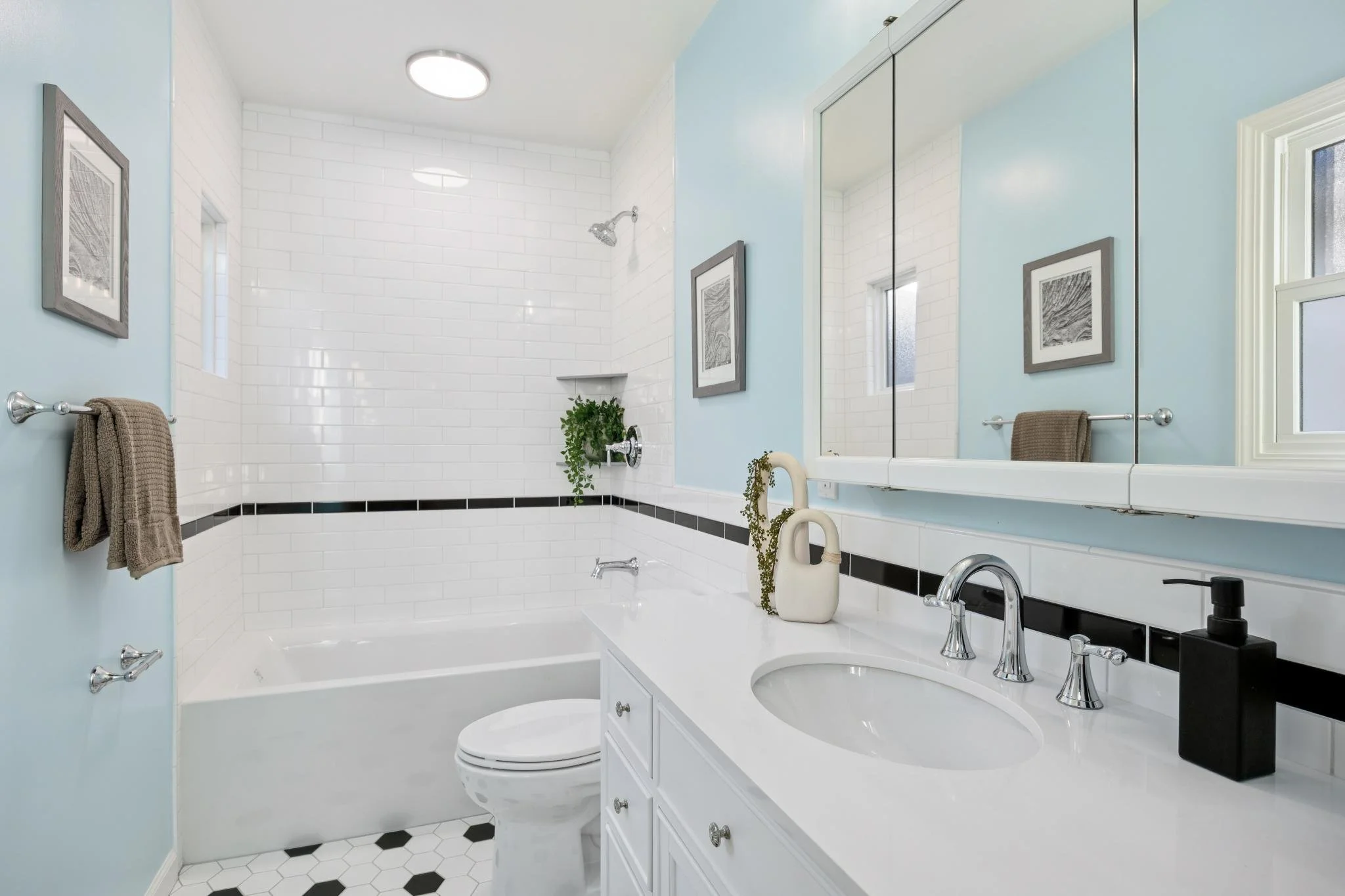 A bright bathroom featuring a white vanity with a marble countertop, a sink, and a black soap dispenser. There is a large mirror above the vanity, a toilet, and a bathtub with a shower. The walls are painted light blue, decorated with framed artwork.