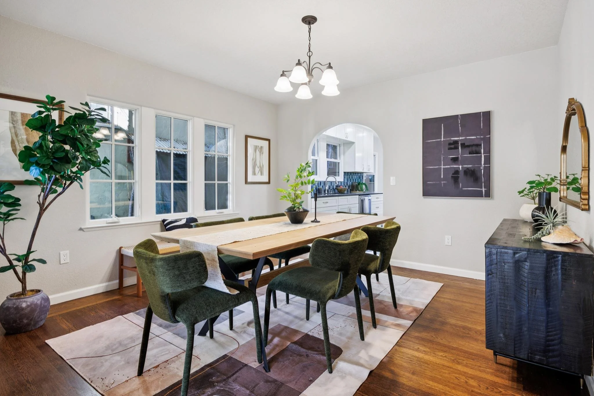 A dining room with a wooden table surrounded by six green upholstered chairs, a chandelier hanging from the ceiling, large windows, and artwork on the walls, with a view of a kitchen in the background.