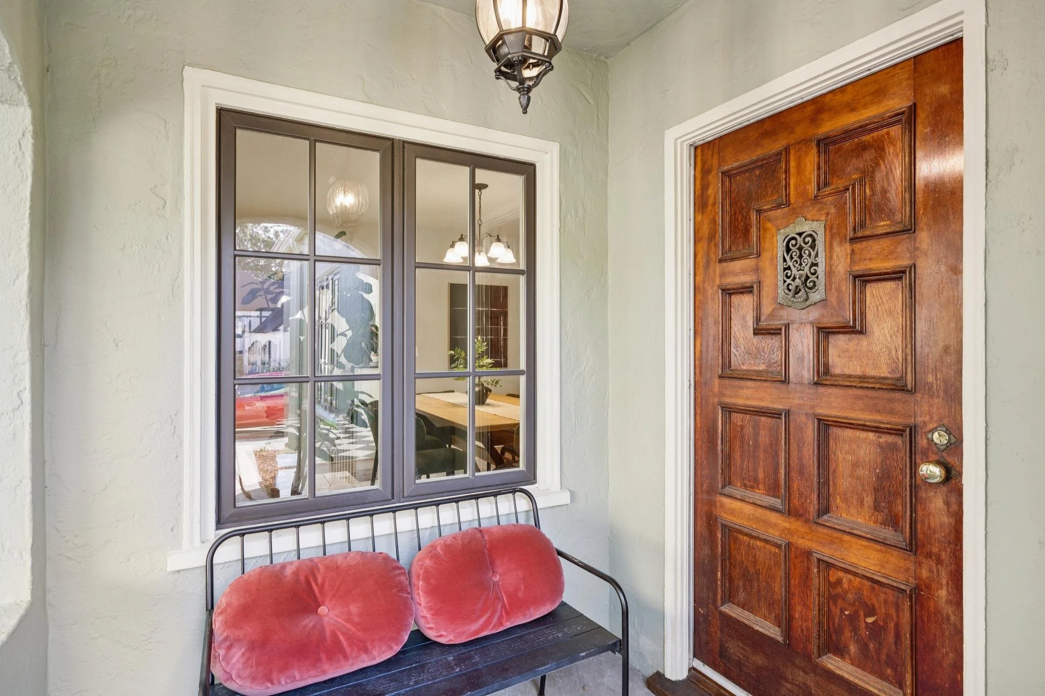 An outdoor porch area with a black metal bench holding red cushioned seats, a wooden front door with decorative metalwork, a window with a gray frame, and a hanging lantern light fixture.
