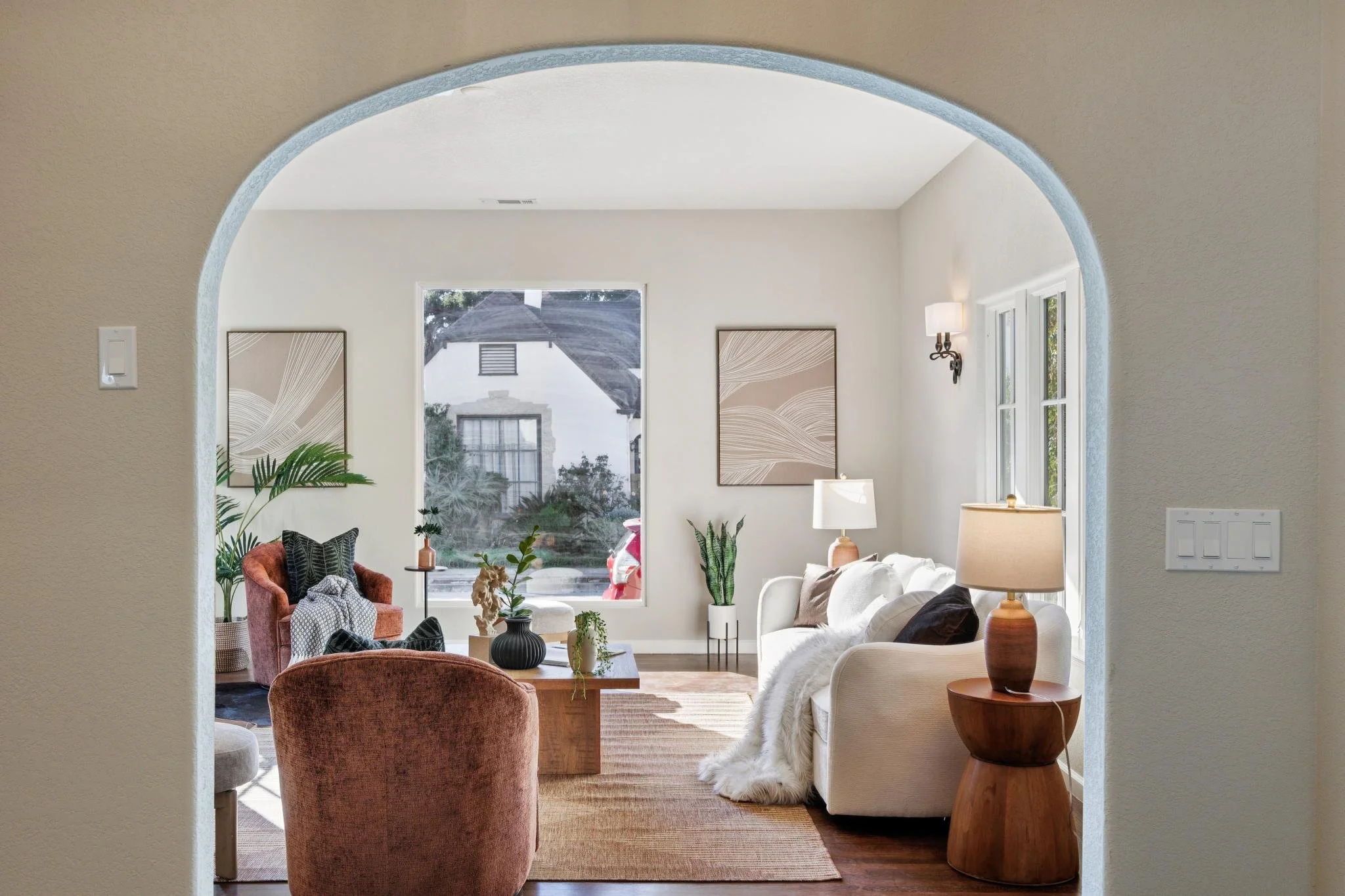 Living room with white walls, large window, white and beige furniture, decorative plants, framed artwork, and a mix of modern and vintage decor.