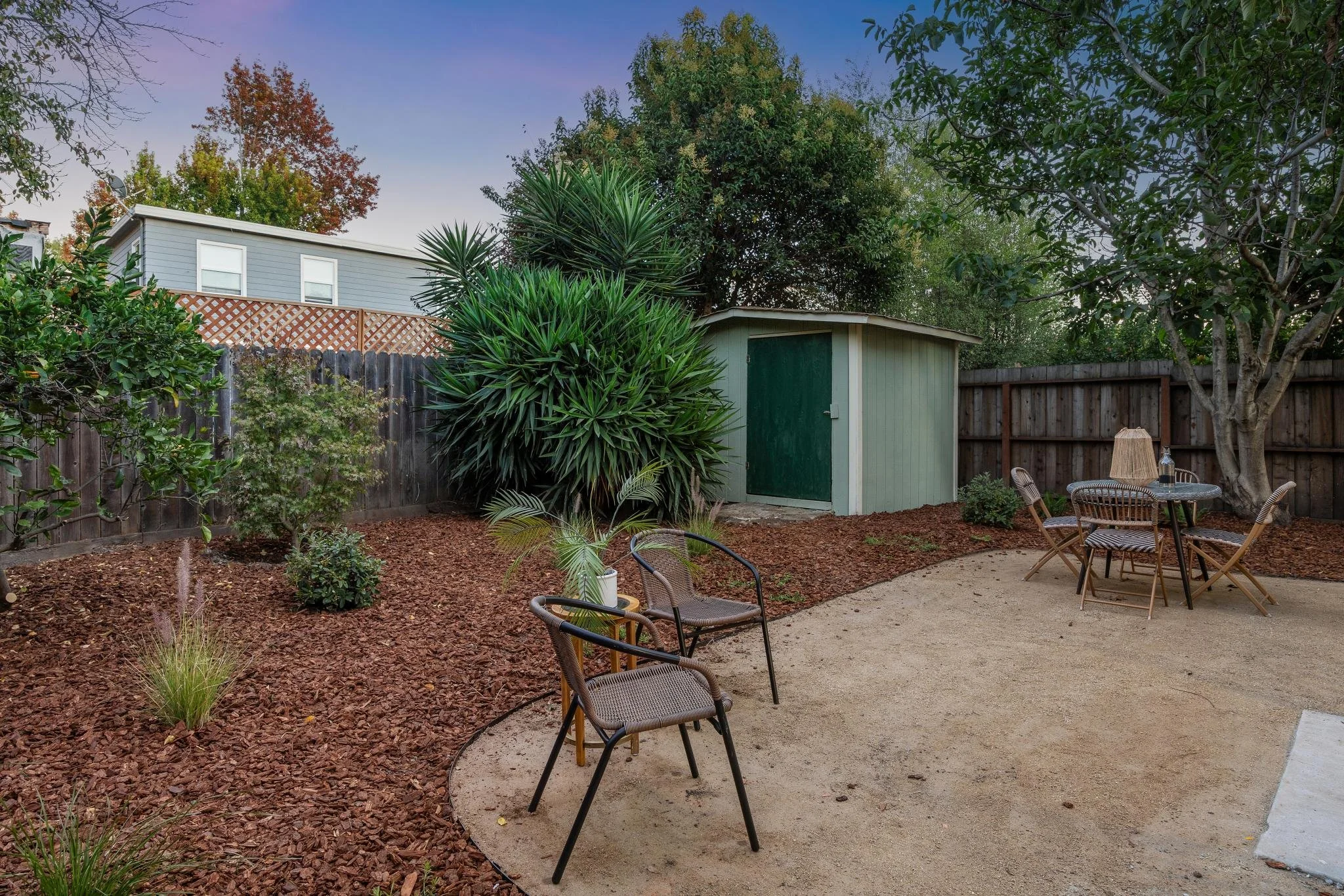 A backyard patio with a circular table and six chairs, surrounded by trees and shrubs, with a small shed and a fence in the background, and a two-story house visible behind the trees.