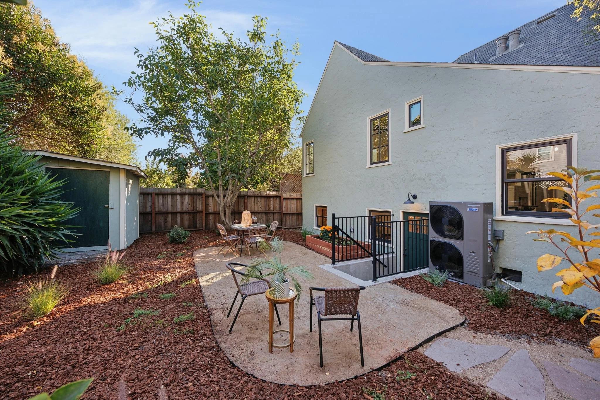 Backyard patio with paved walkway, outdoor furniture, lush plants, a small tree, a shed, a tall hedge, and a pale green house with multiple windows and outdoor heating units.