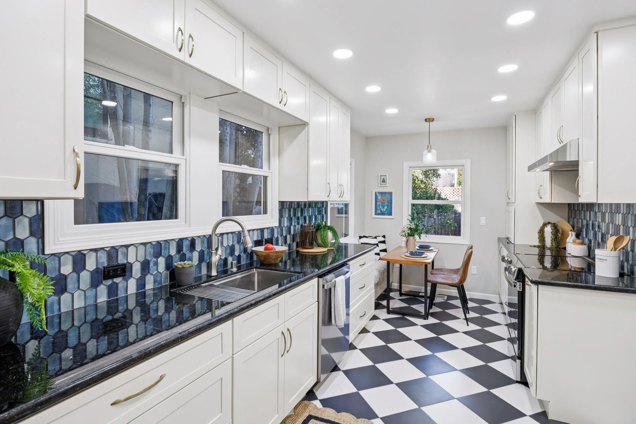 A modern kitchen with white cabinets, blue hexagonal tile backsplash, black countertops, black and white checkered floor, and large windows. There is a dining table with chairs, plants, and artwork on the wall. Recessed lighting and a hanging light fixture are visible.