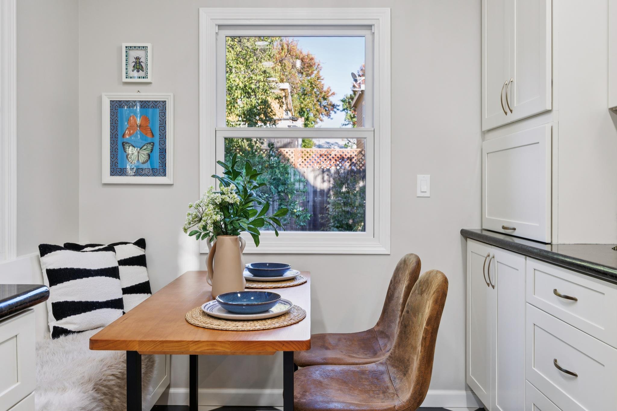 A cozy dining area with a wooden table set with three plates on woven placemats, a beige vase with green foliage, and two chairs. The space features a window showing an outdoor scene with trees and rooftops. The wall decor includes framed butterfly a