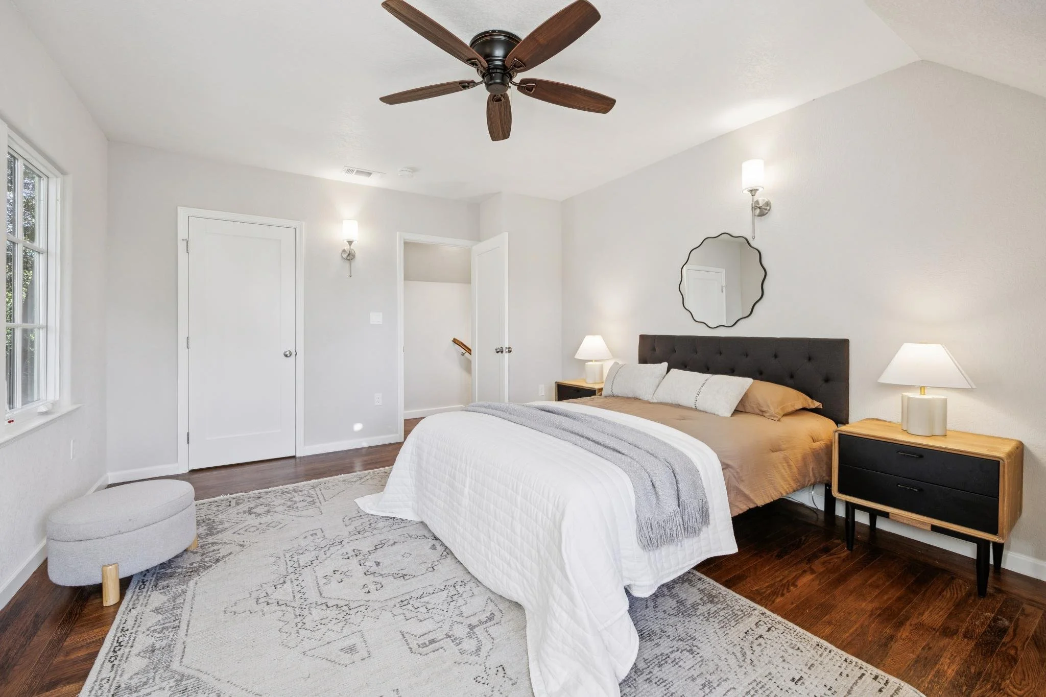 A bedroom with a large bed covered in white bedding, a beige blanket, and multiple pillows. There are two wooden nightstands with lamps on either side of the bed. A wall mirror is mounted above the headboard, and a ceiling fan with wooden blades is i