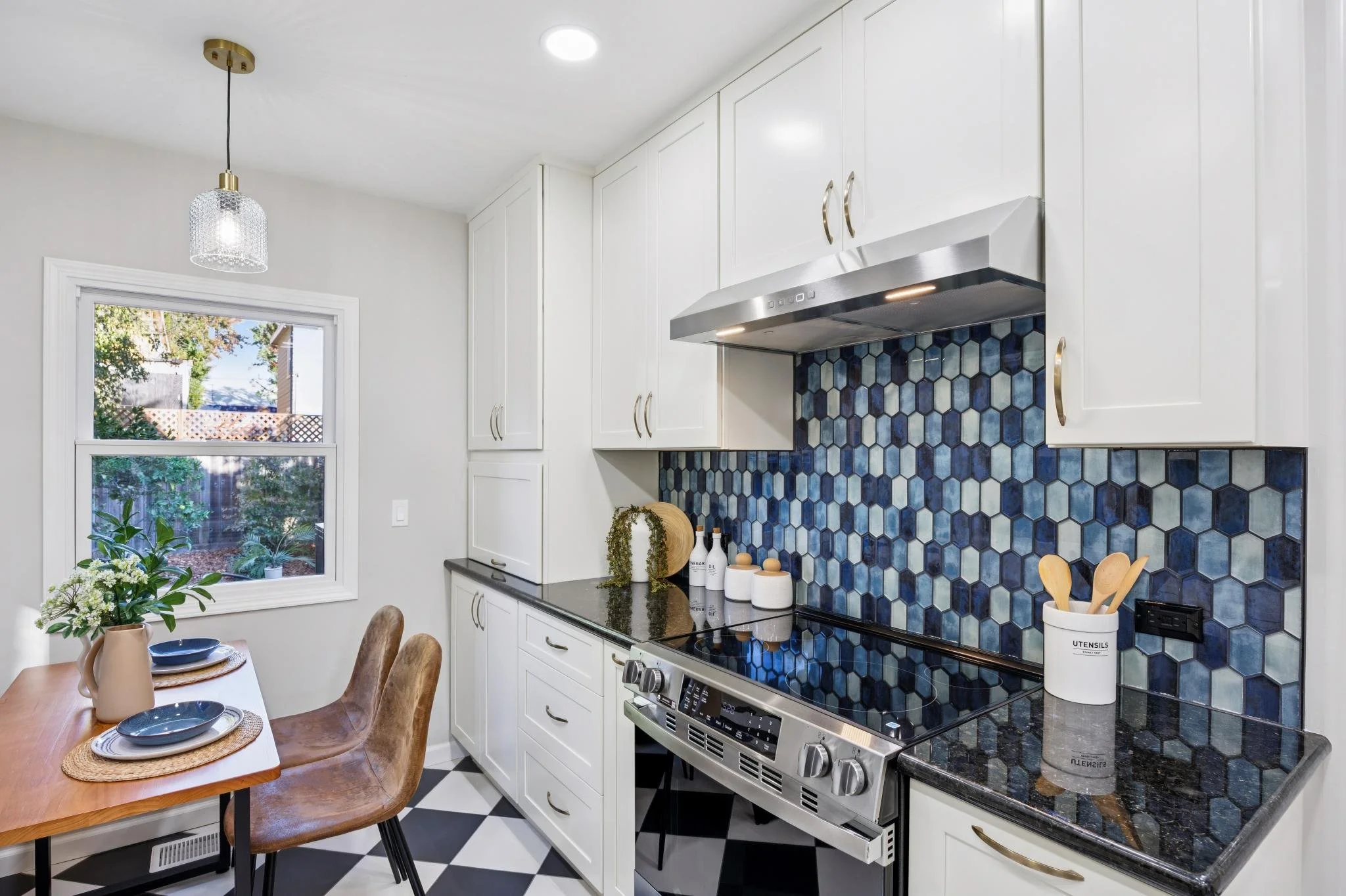 A kitchen with white cabinets, a black and blue hexagonal tile backsplash, stainless steel appliances, a window overlooking a garden, a dining table with place settings, and decorative vases with plants.