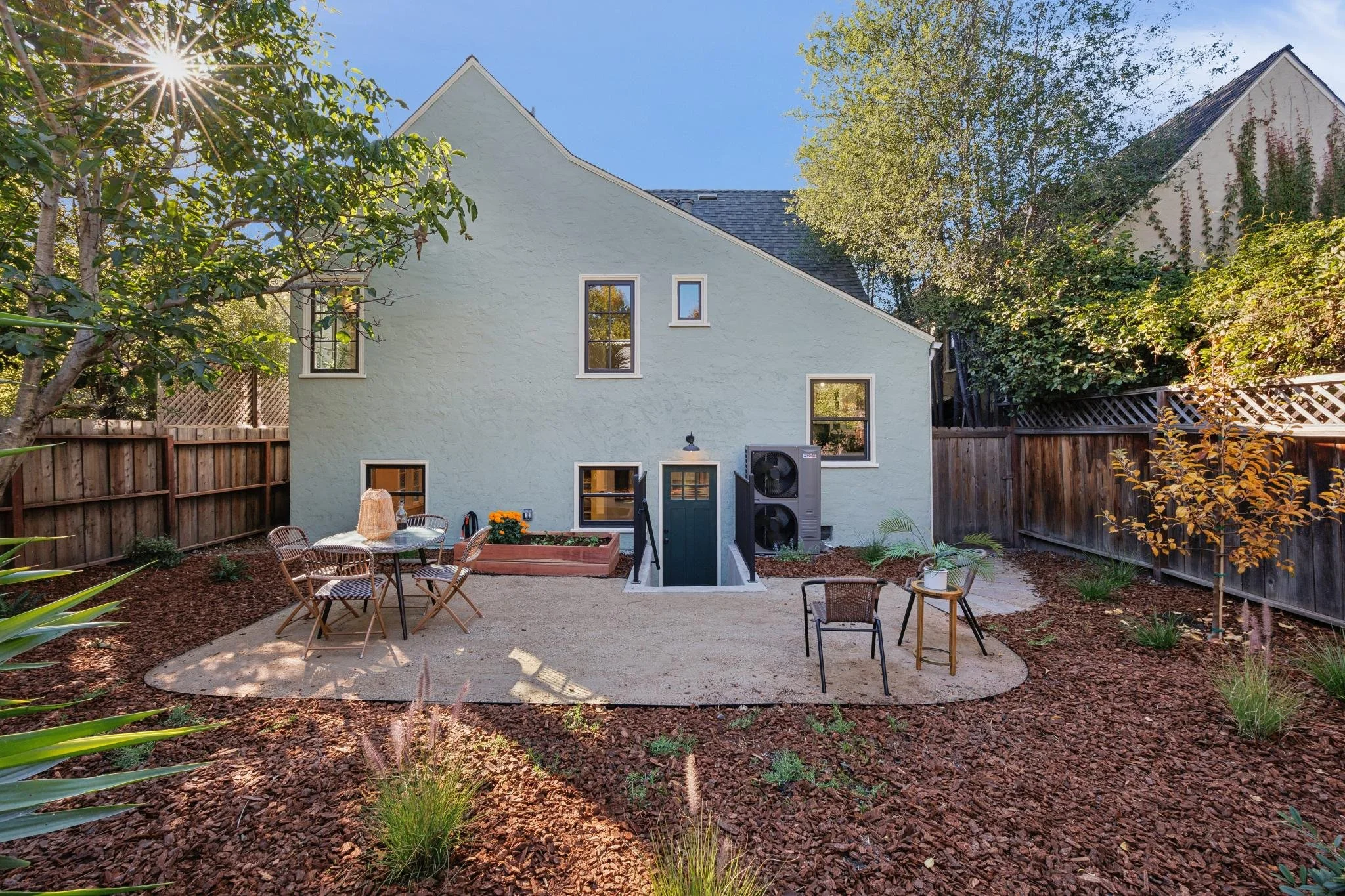 Backyard with wooden fence, patio furniture, small trees, potted plants, and a house with three windows and an HVAC unit outside, under a clear blue sky with the sun shining.