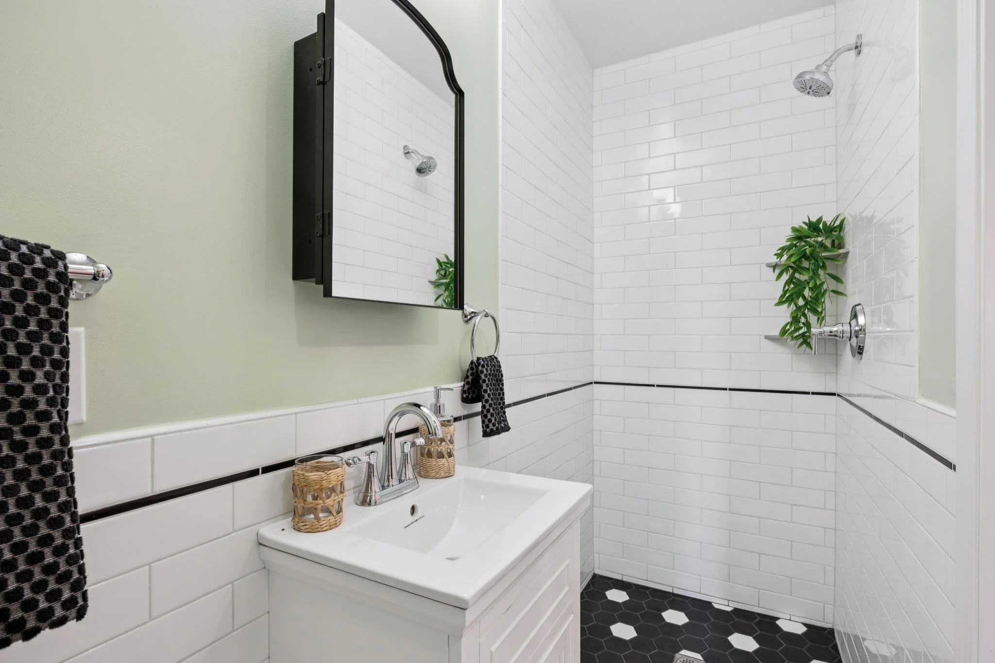 A bathroom with a white vanity, black and white hexagonal tile floor, and a shower with white subway tiles. There are black patterned towels hanging on a towel rack and a mirror on the wall, with a plant placed on the shower ledge.