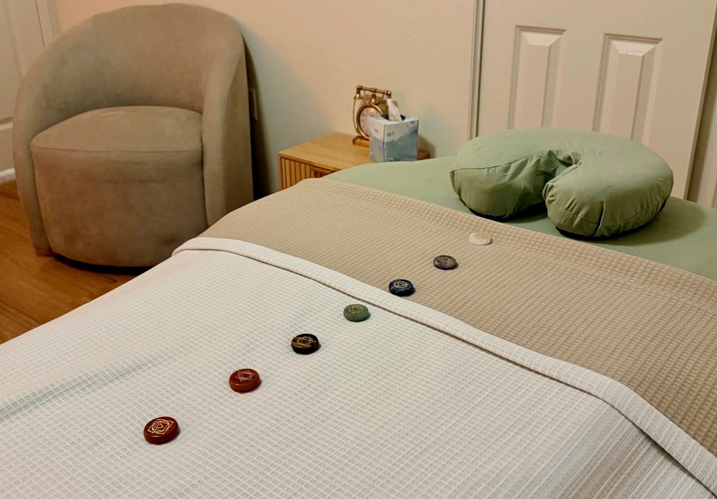 A bed with a green circular pillow and a series of colorful stones placed in a line on top of a white textured blanket. A beige armchair is beside a wooden nightstand, which holds a small floral clock and a tissue box. A white door is in the background.