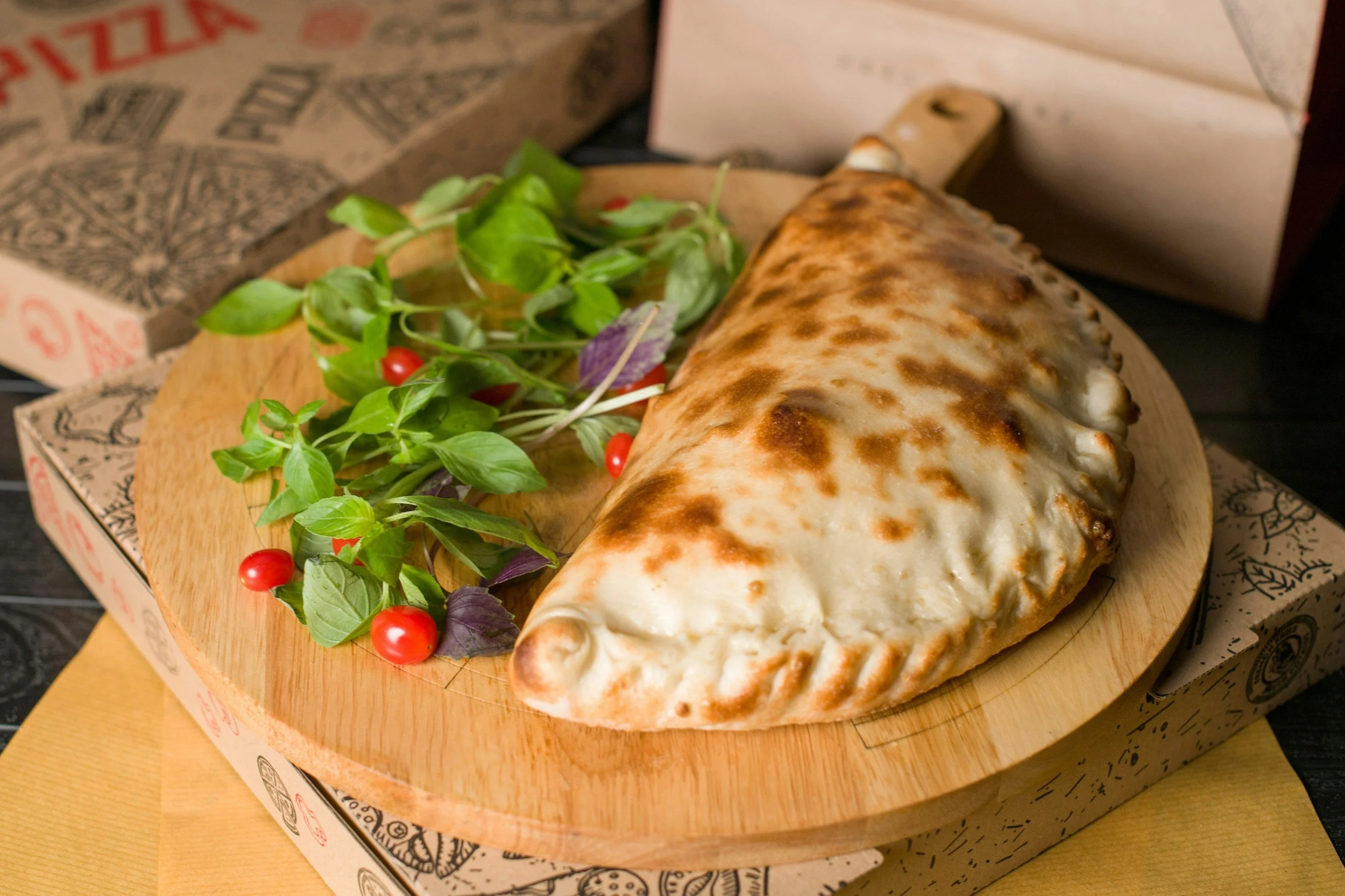 Baked calzone with a golden-brown crust on a wooden serving board, garnished with a side of fresh green and purple basil leaves and red cherry tomatoes, with pizza boxes in the background.