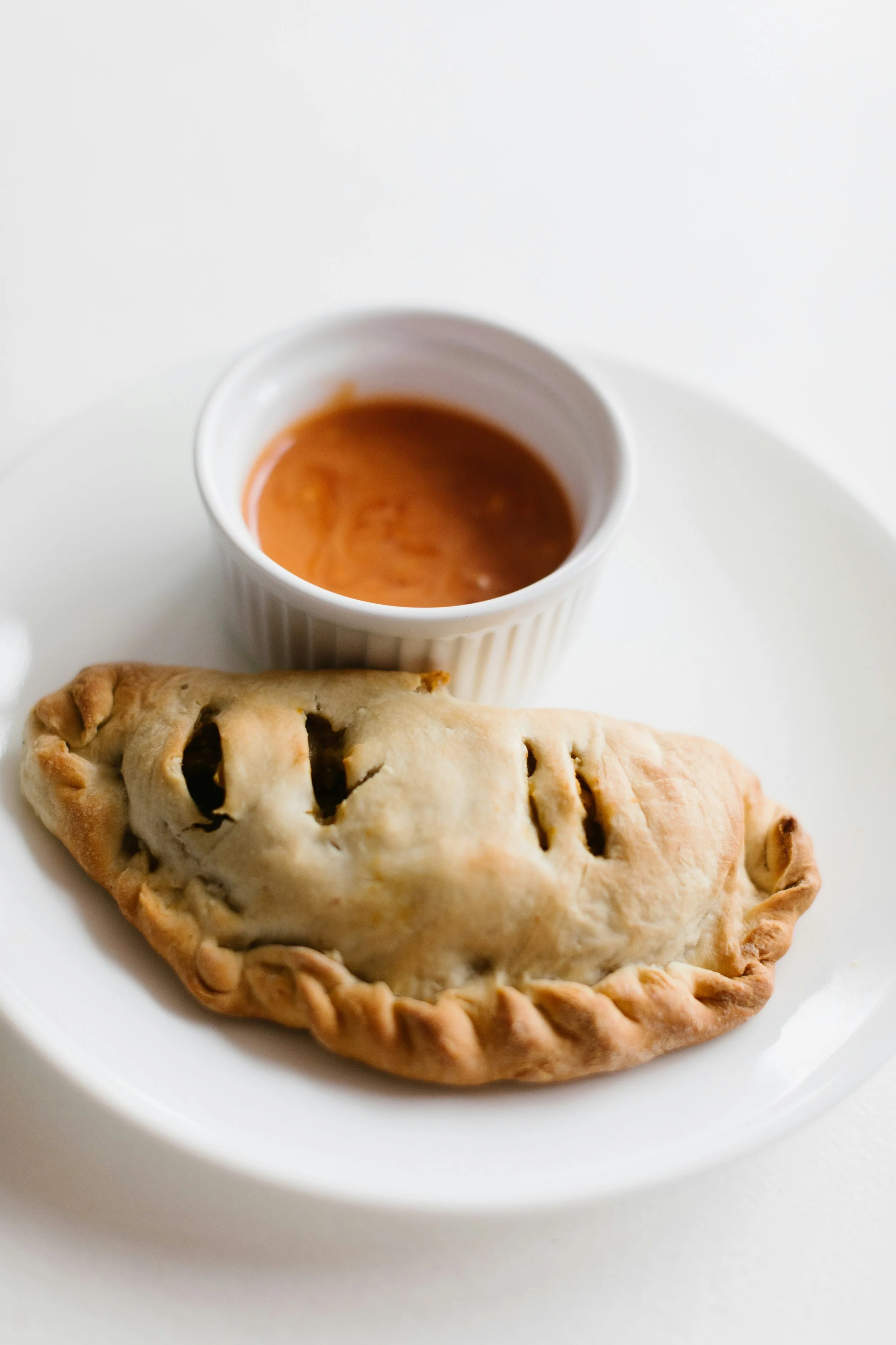 A white plate with an empanada and a small bowl of dipping sauce on a white surface