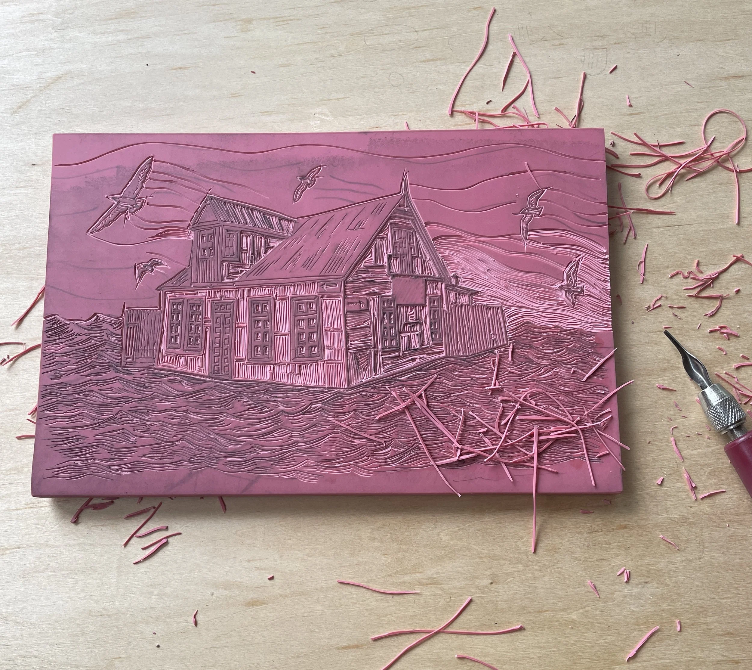Pink cutting board with a detailed engraving of a house and seagulls, with pink rubber strips and scissors around it.