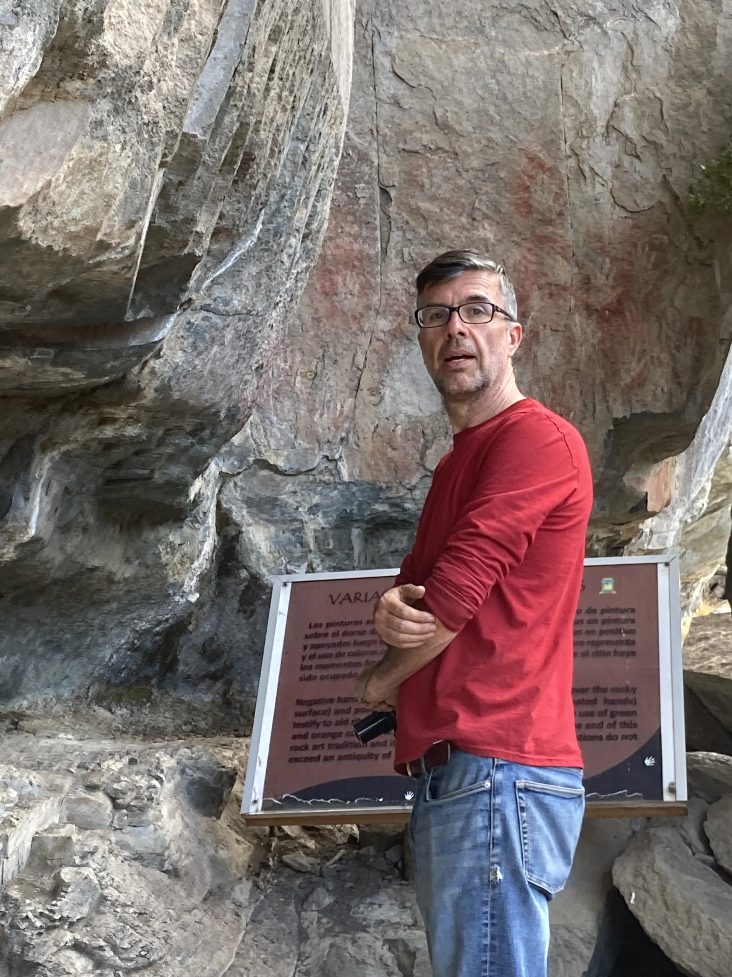 A man with glasses and a red shirt standing in front of a rock wall and an informational sign.