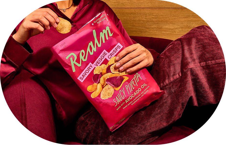 A woman in a deep red shirt and matching corduroy pants lounges on a red chair and holds Realm Saucy Pizza Chips packaging.