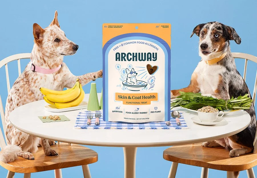 Two mixed-breed dogs sit at a table with a package of Arch Dog Treats.