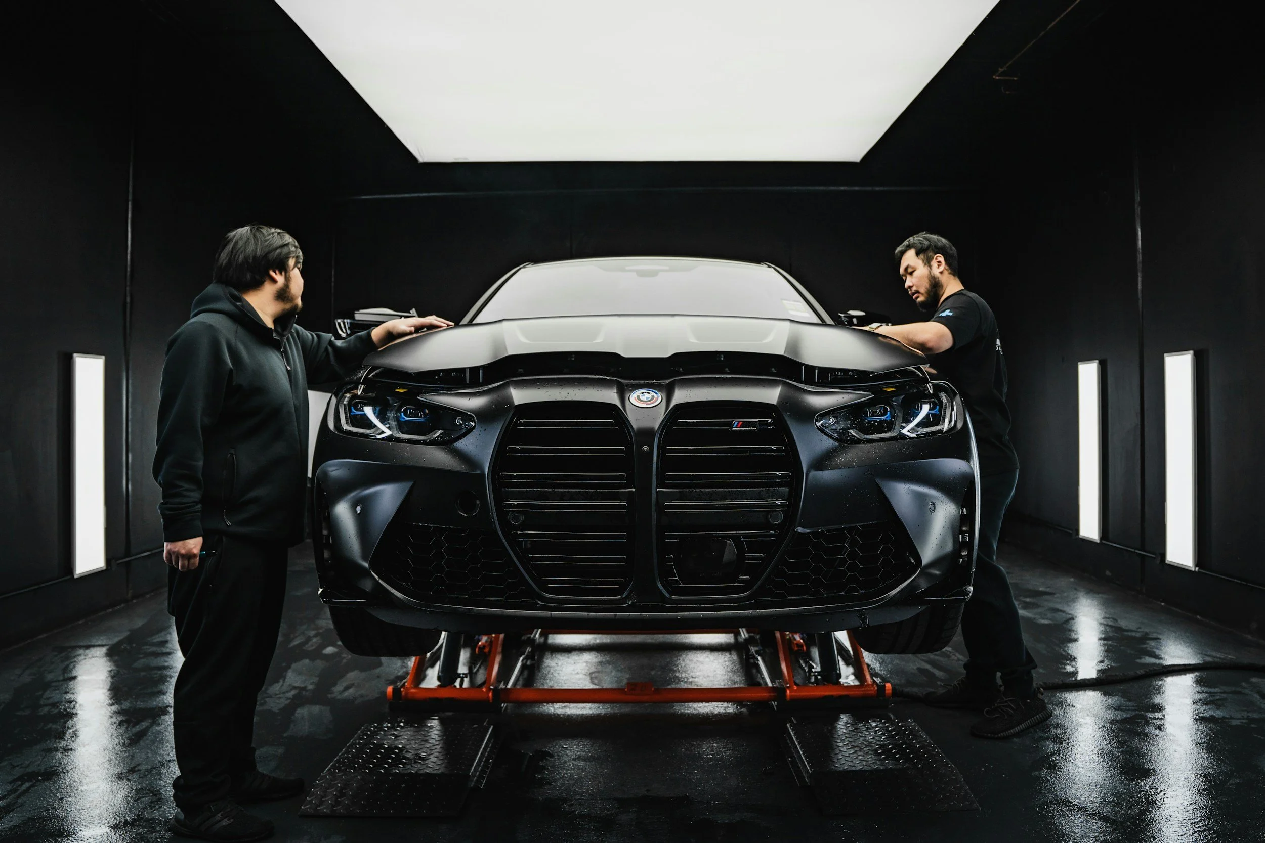 Two men work on a black sports car inside a dark garage with a white ceiling light, adjusting or inspecting the front of the vehicle.
