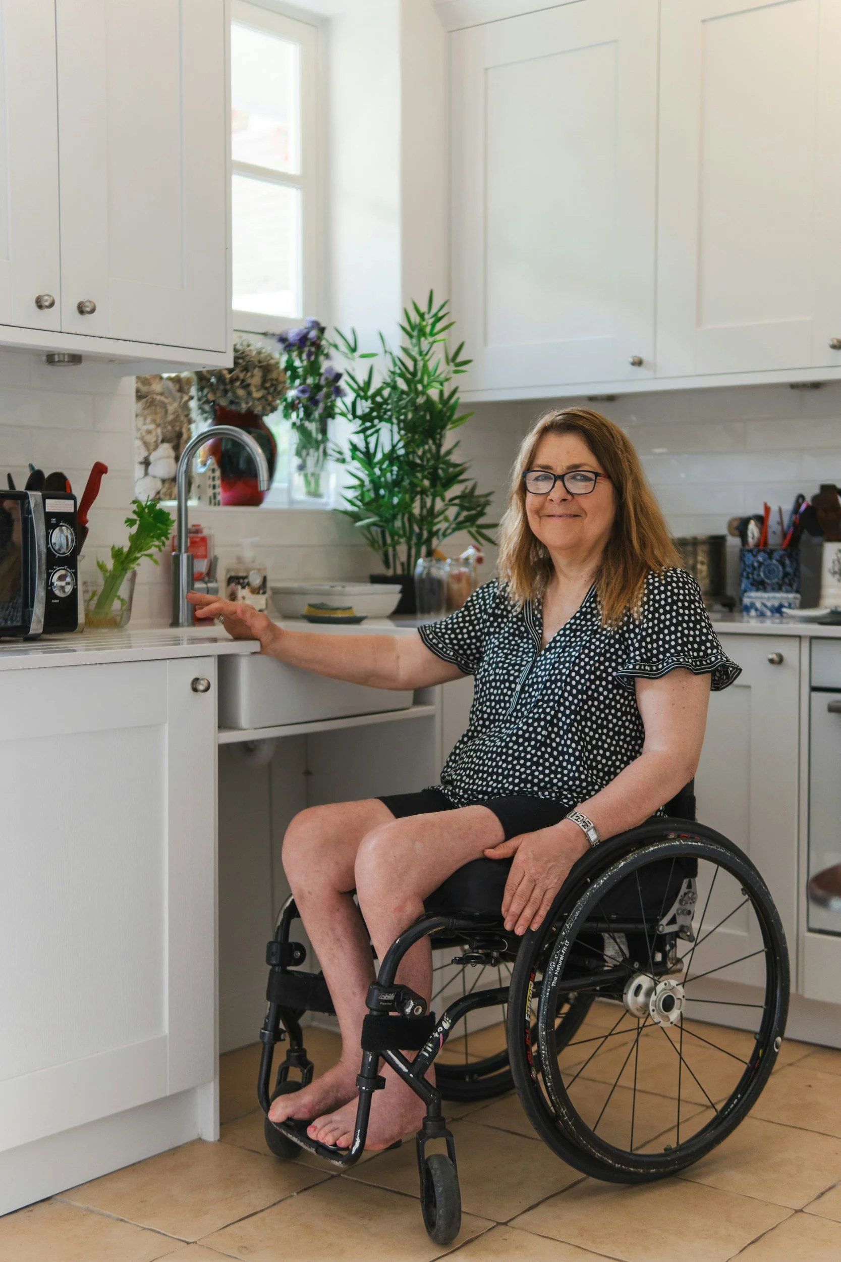 A woman with glasses in a wheelchair in a bright kitchen, smiling, wearing a black and white polka-dot blouse, with green plants and kitchen appliances in the background.