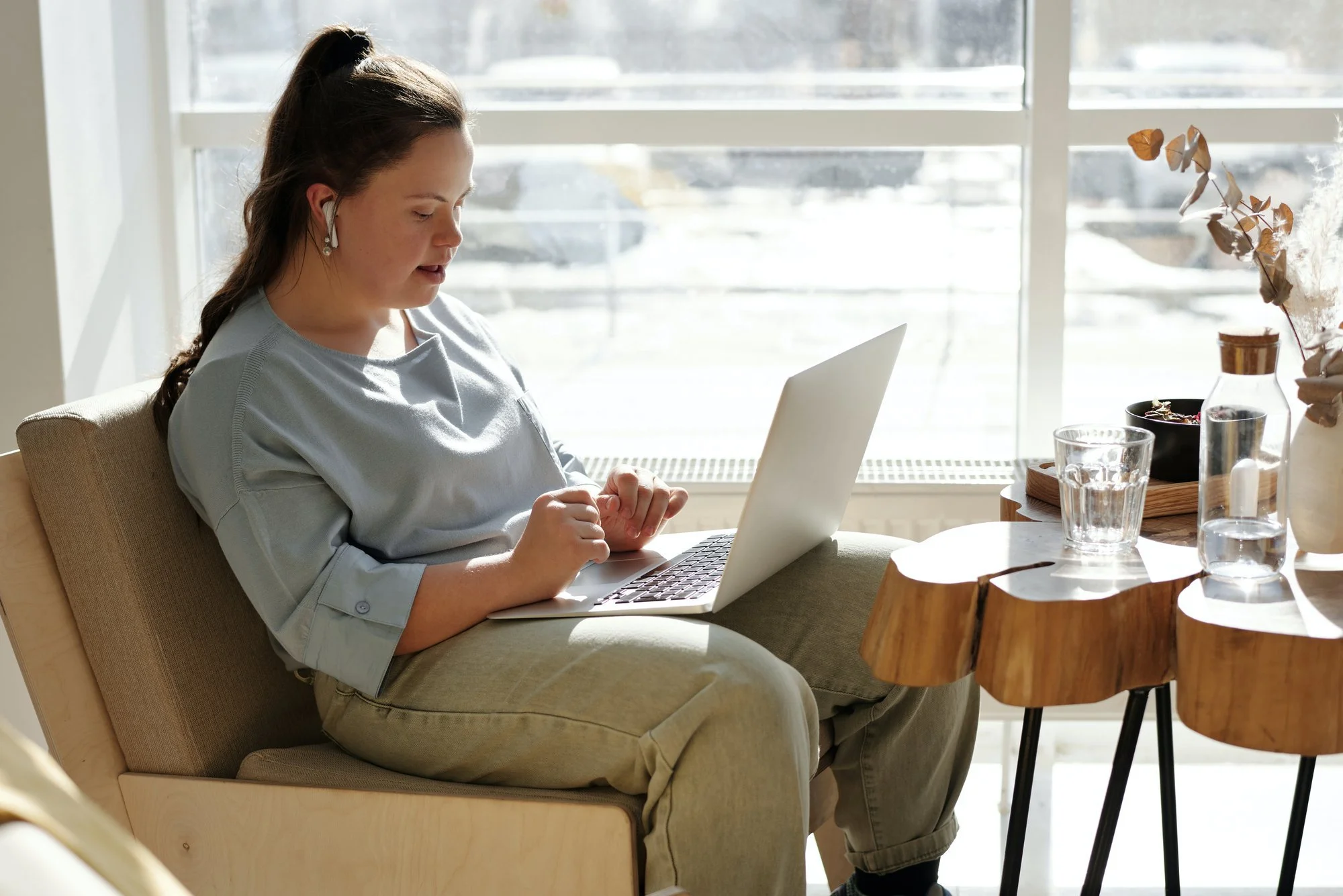A woman sitting on a beige chair at a table, working on a silver laptop. She is wearing a light blue top and beige pants, with earbuds in her ears. Sunlight streams through large windows behind her, illuminating the room. On the table, there are glass cups, a small wooden tray, and decorative dried plants in vases.