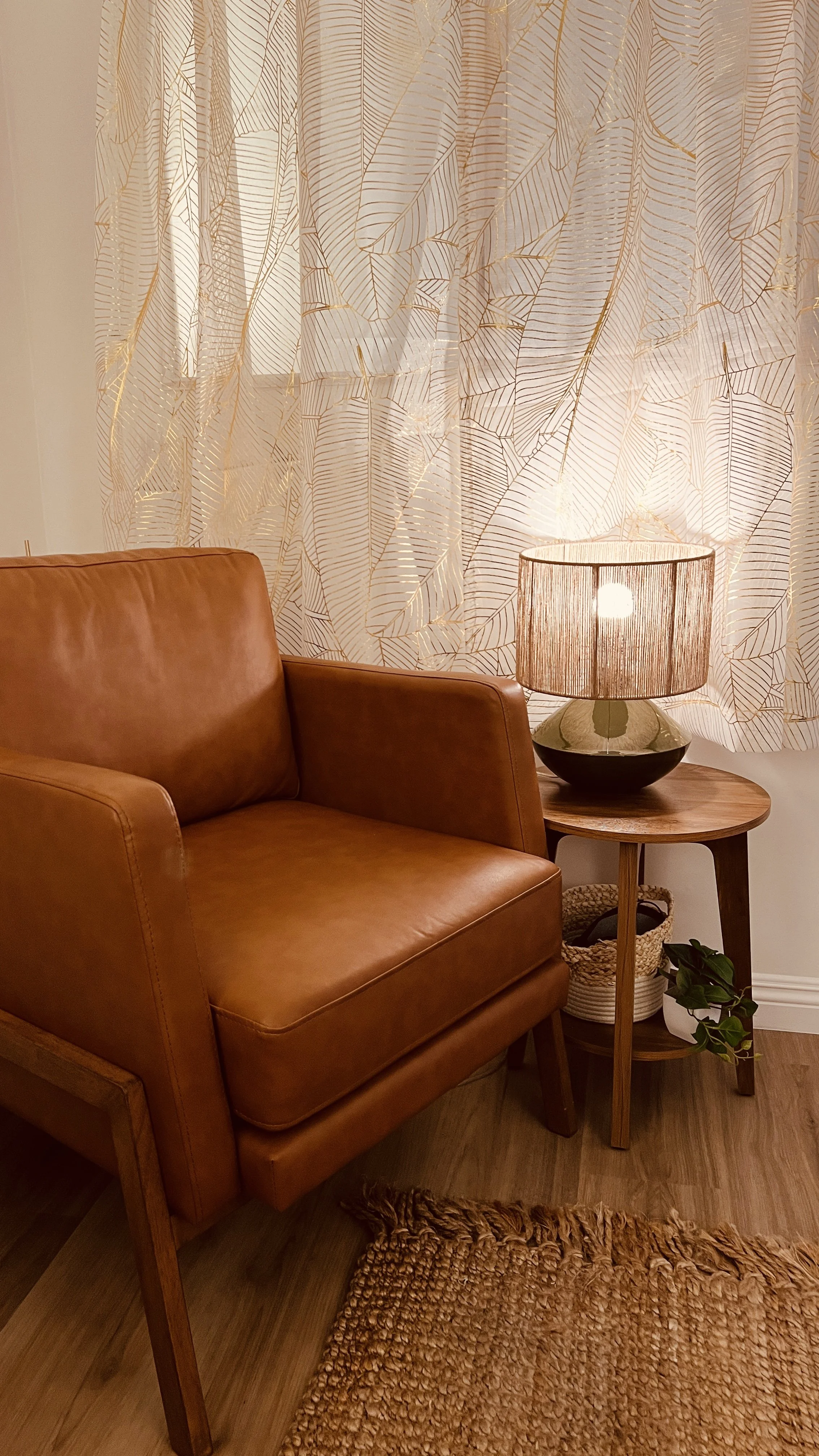 A cozy living space corner with a tan leather sofa, a wooden side table, a table lamp with a beige shade, a woven basket, and a potted plant on the floor, with patterned curtains in the background.