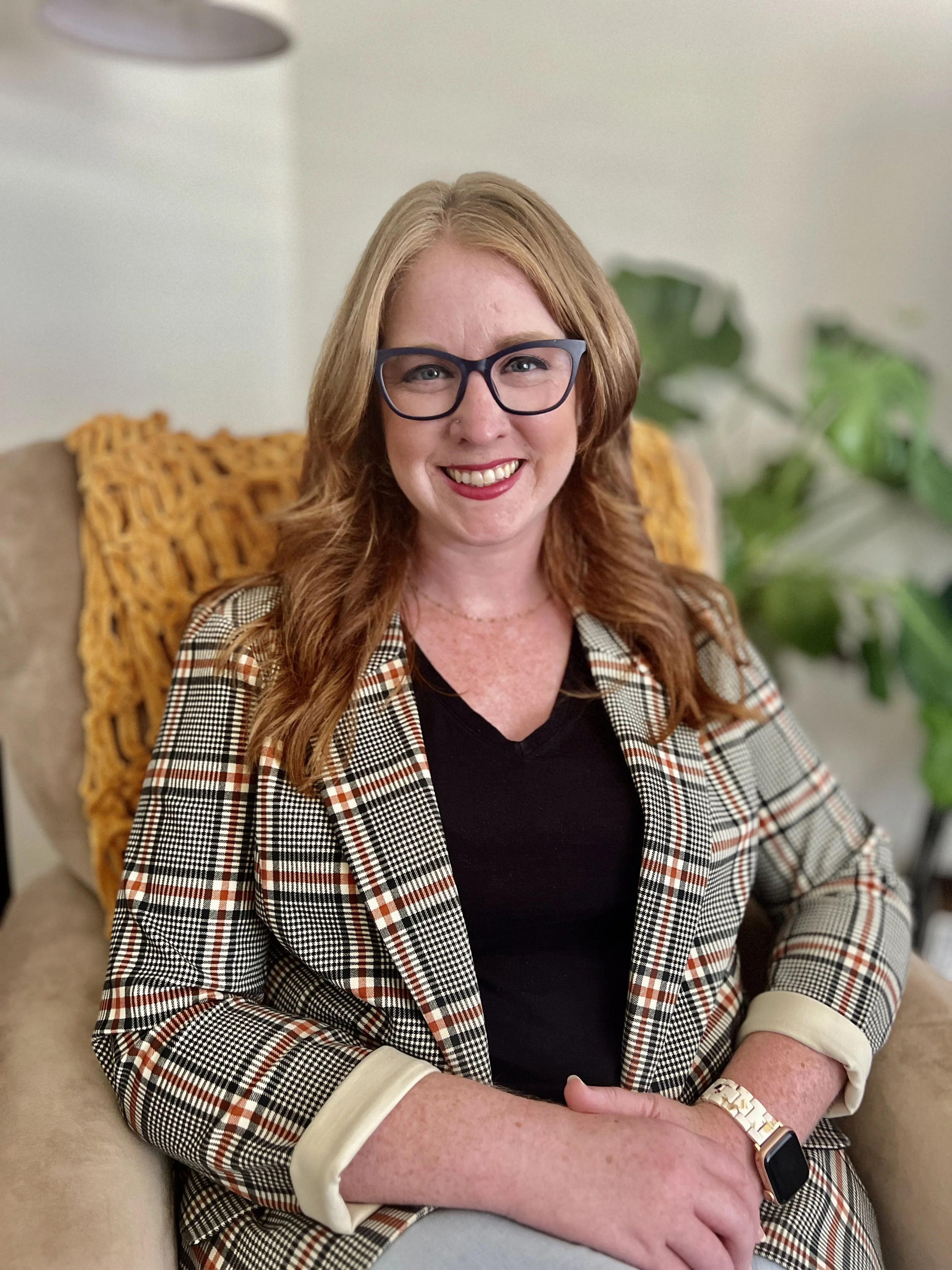 A woman with red hair, glasses, and a checkered blazer smiling while sitting on a beige armchair with a yellow knitted pillow behind her. She is wearing a black top, a gold watch, and a smartwatch on her left wrist. There are green leafy plants in the background.