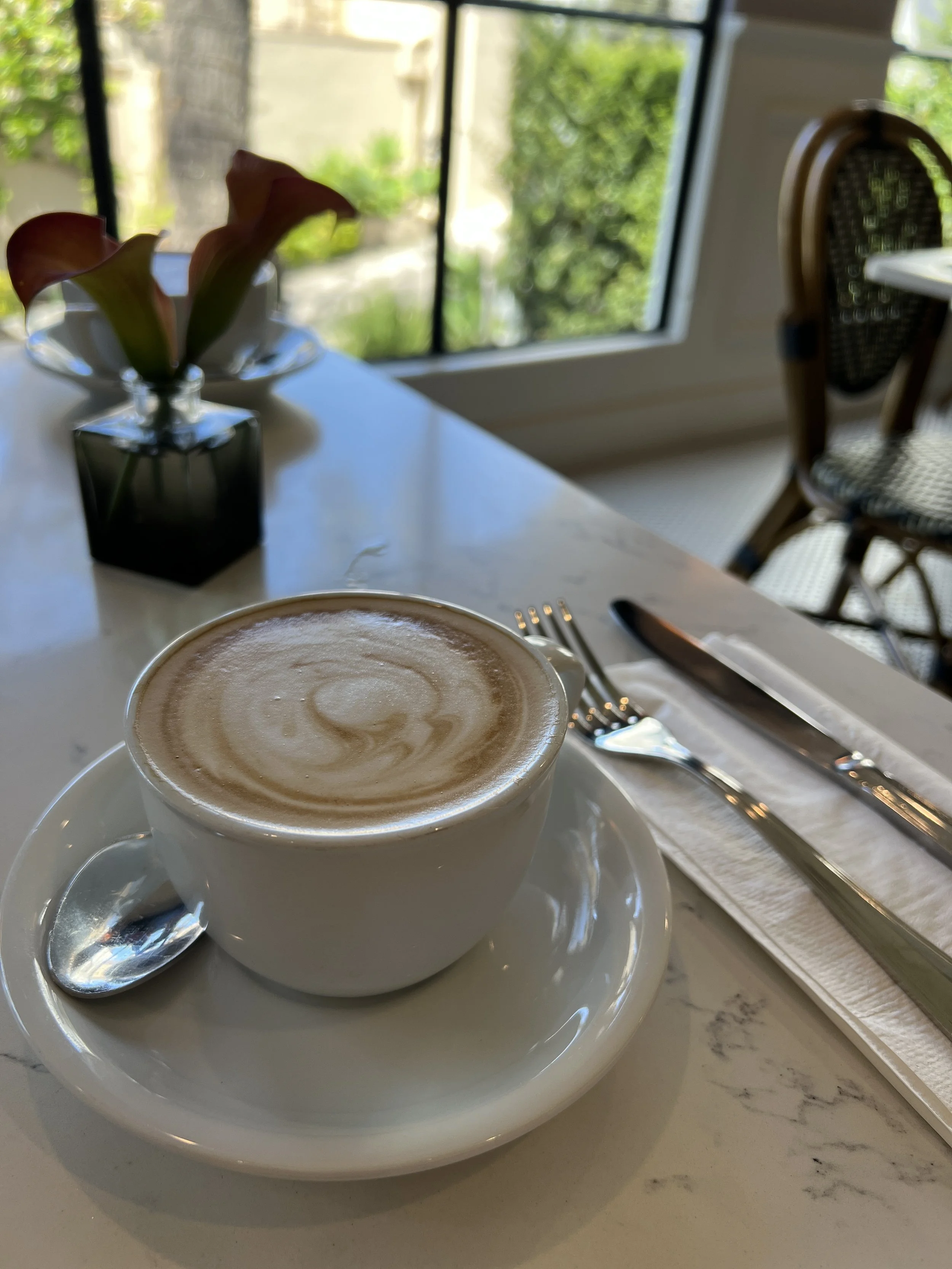 A cup of coffee with foam art on a table at a cafe, with a flower in a vase and a window showcasing greenery outside.