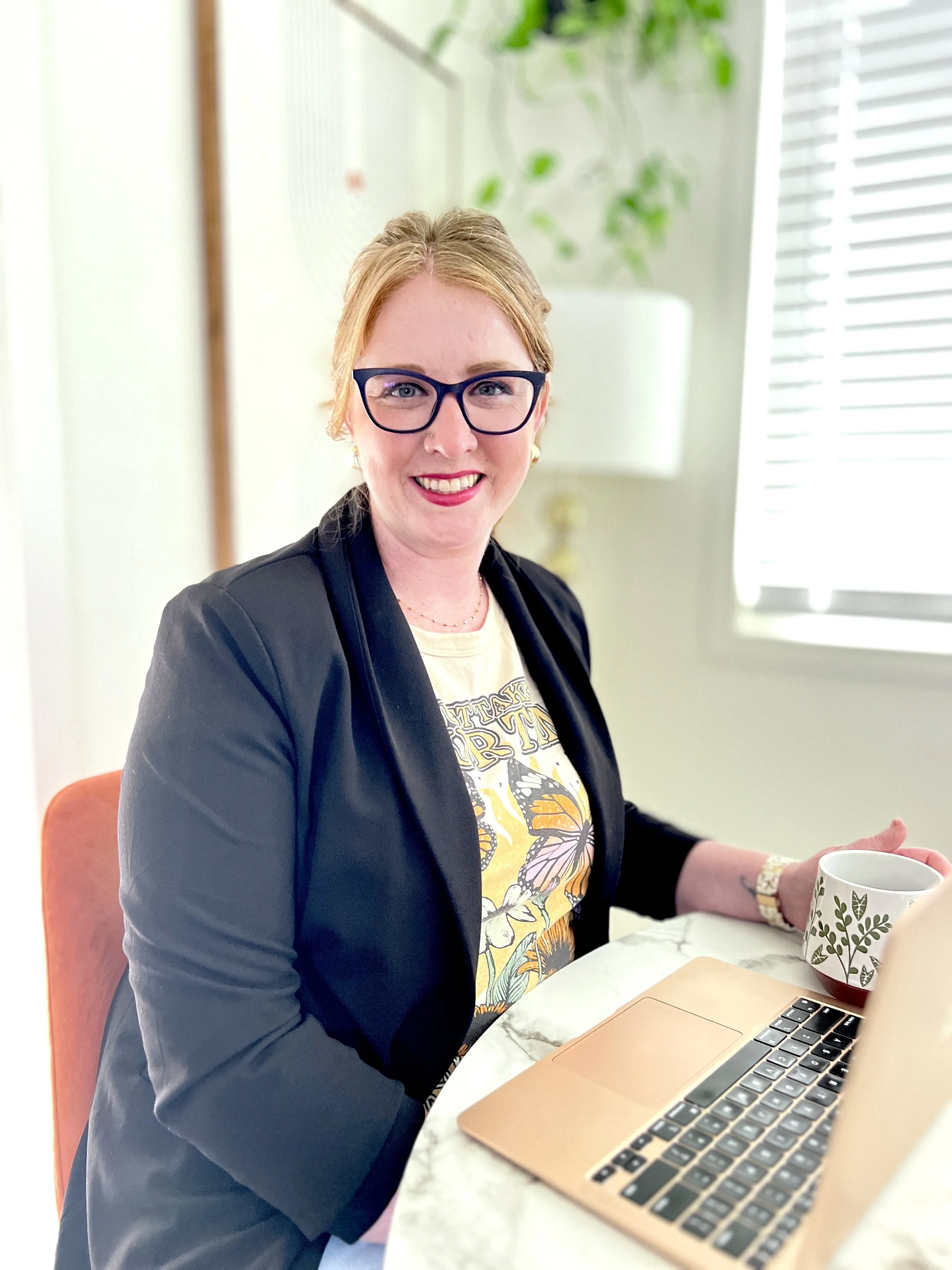 A woman with blonde hair, glasses, and a black blazer sitting at a table with a laptop and a coffee mug, smiling at the camera in a bright room with blinds and a potted plant in the background.
