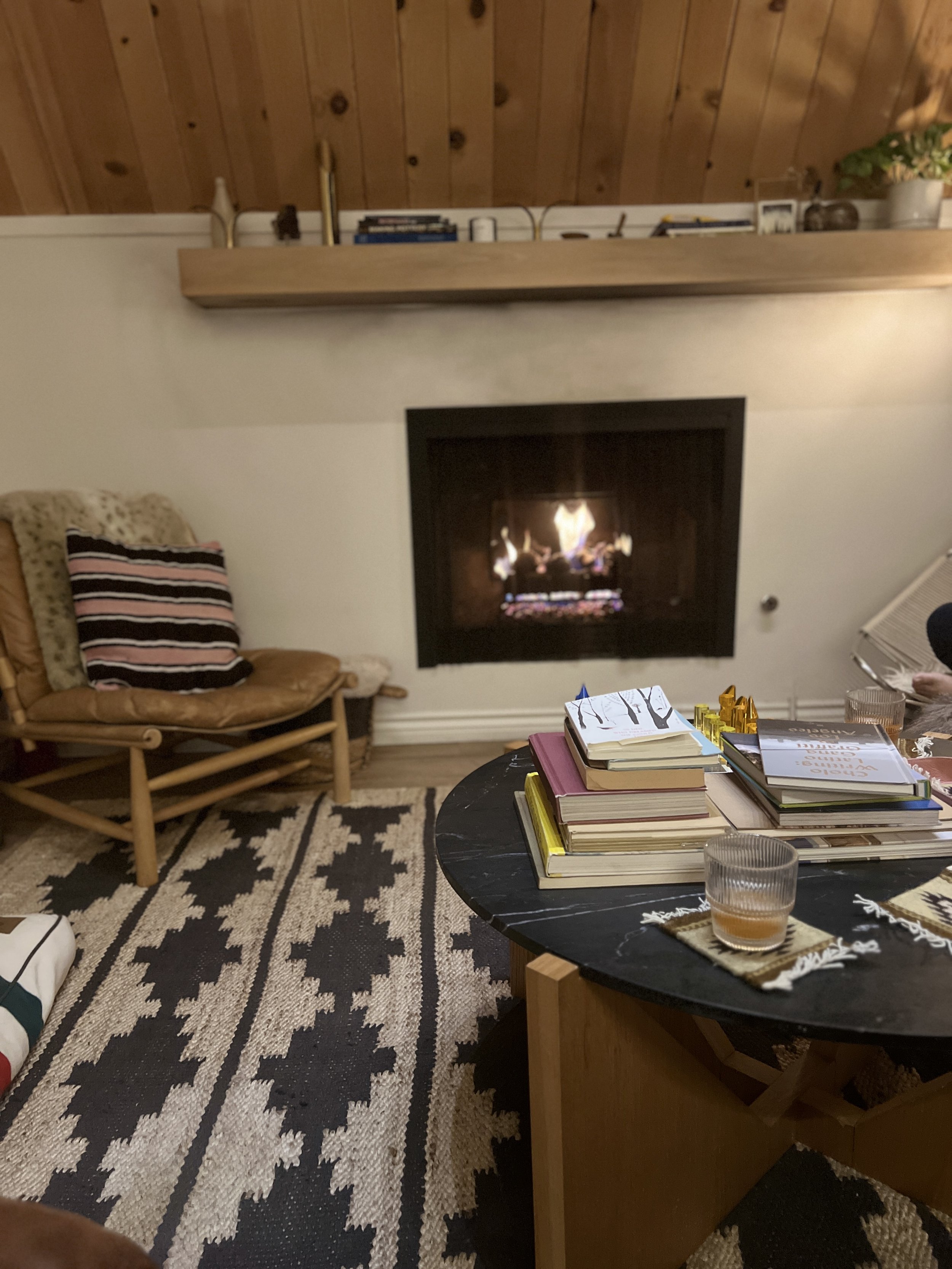 Living room with wood-paneled ceiling, fireplace with a fire, small wooden chair with a leopard-patterned cushion, black and white rug, black marble coffee table with books and drinks, and a stack of books on a side table.